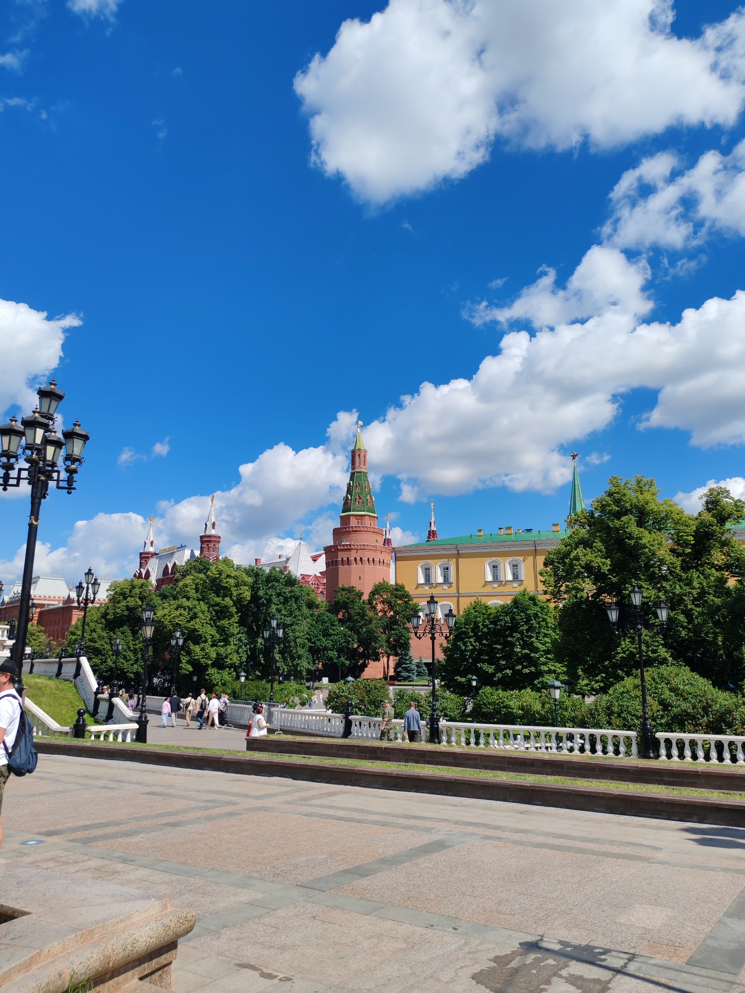 The image shows a sunny day in Moscow, Russia, with a clear blue sky filled with fluffy white clouds. In the background, the iconic red brick walls and green-domed towers of the Moscow Kremlin are visible, along with a yellow building that appears to be part of the complex. Lush green trees are scattered throughout the scene, providing shade and contrast to the historic architecture.
In the foreground, a paved walkway leads to a grassy area with a white balustrade. Several people are walking and standing in this area, enjoying the pleasant weather and the scenic views. A black lamppost with multiple lights stands tall on the left side of the image, adding to the classic European aesthetic. The overall atmosphere is one of a lively and beautiful day in a historic city.