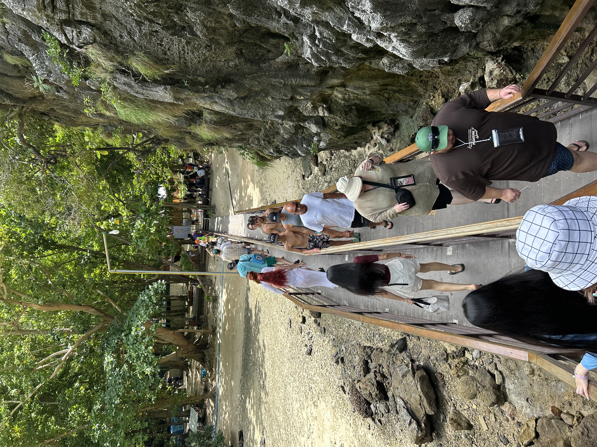 A group of tourists walk along a wooden boardwalk through a rocky, vegetated area near a beach in Ao Nang, Thailand. The boardwalk is elevated and lined with a wooden railing. On one side of the boardwalk is a sandy beach with trees and some buildings, possibly shops or restaurants. On the other side, a sheer rock face covered in moss and grass rises steeply. The tourists are dressed in casual, warm-weather clothing, and some are carrying bags. The scene appears to be a popular tourist attraction, with many people present. The weather is sunny and bright, suggesting a warm day.