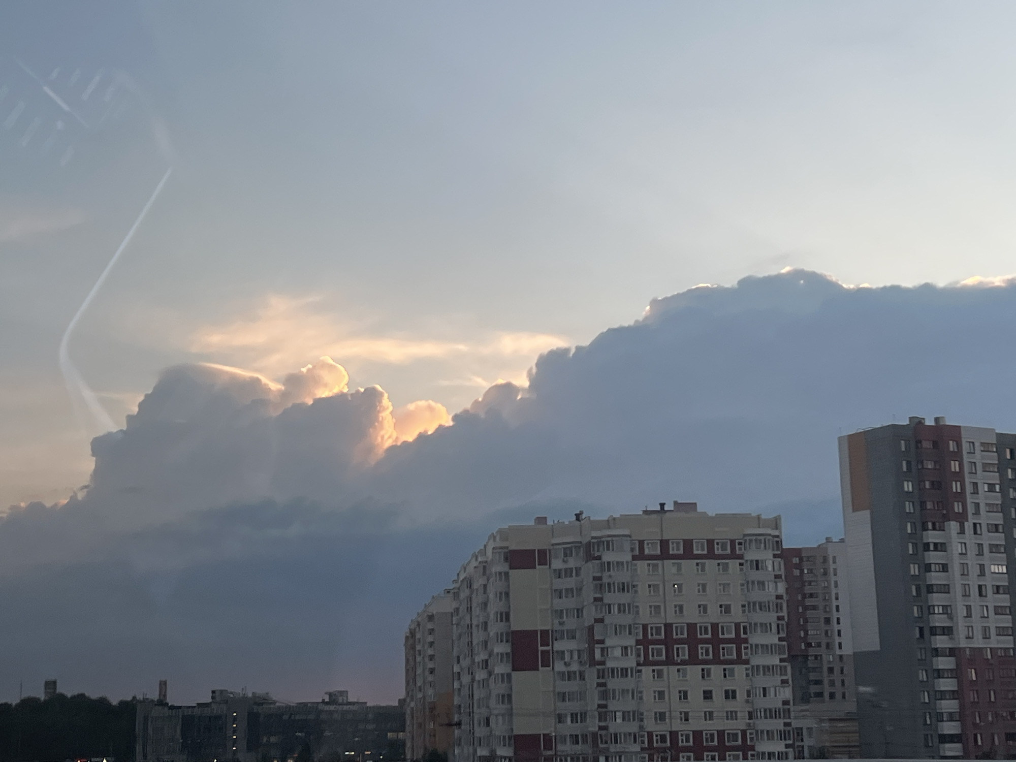 The image shows a cityscape under a cloudy sky. The clouds are dense, with sunlight peeking through some areas, creating a dramatic lighting effect. In the foreground, there are several high-rise apartment buildings. In the distance, there is a row of smaller buildings, possibly within a forested area. The location is unknown.