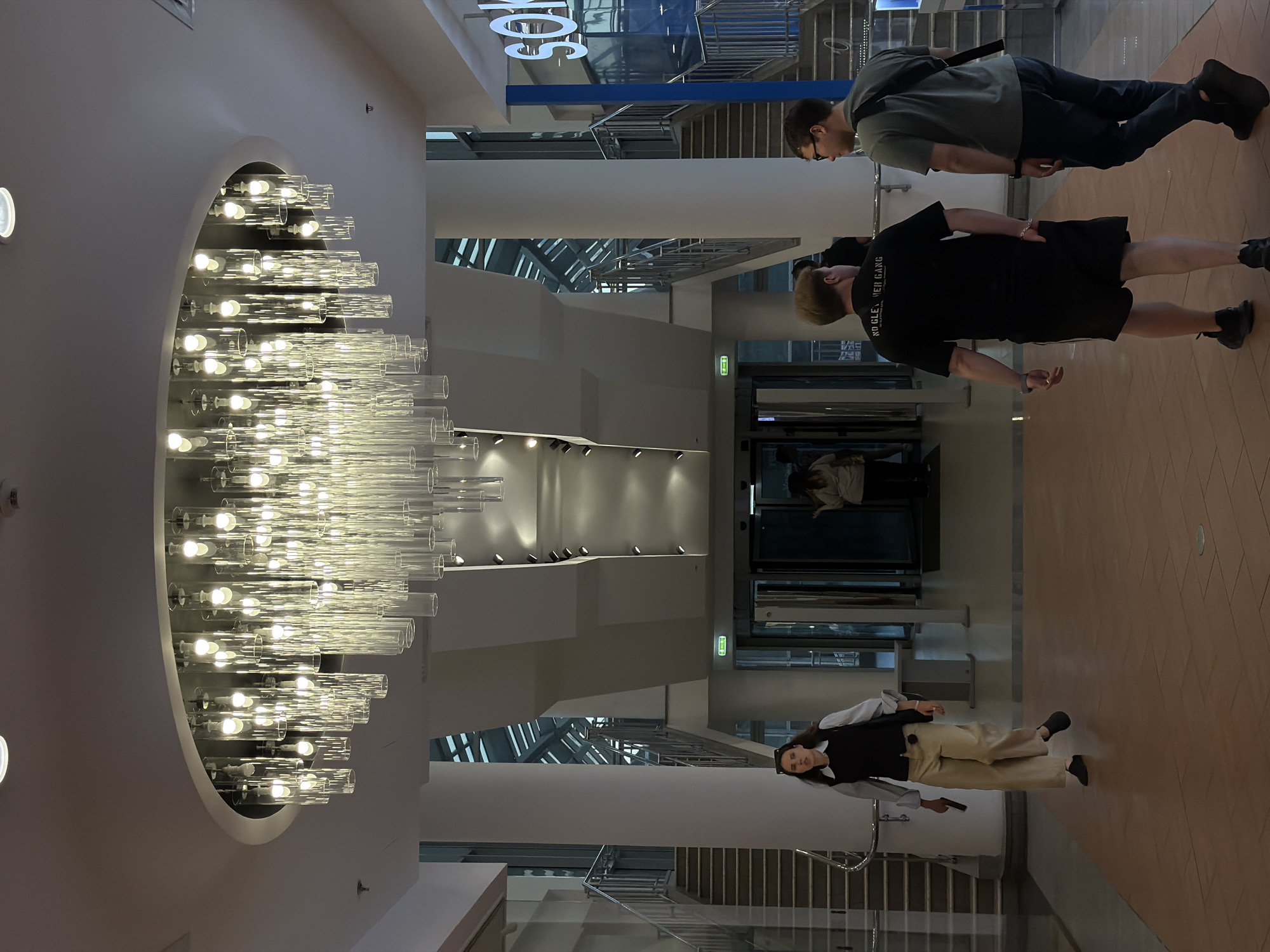 This image depicts an interior public space, likely a multi-level shopping mall or large atrium, located in Moscow, Russia. The perspective is from the ground floor, looking towards a back area with stairs and glass doors. Dominating the overhead view is a large, modern chandelier composed of numerous illuminated cylindrical glass tubes of varying lengths, suspended from a circular recess in the white ceiling. Additional smaller, recessed spotlights are visible in the ceiling. The architectural elements include two large, white cylindrical columns. One is prominent in the mid-ground, while another is partially visible on the far left. Stairs with metal handrails ascend on both the left and right sides of the space, leading to upper levels. Above these stairs, a multi-story glass roof or skylight structure is visible, allowing natural light into the interior. At the far end, beyond the central column, automatic glass doors are visible, with a green illuminated exit sign above them. The floor is covered with reddish-brown tiles arranged in a pattern, with several small circular metal floor access points or lights embedded in it. Several individuals are present: * In the left foreground, a woman with dark hair, wearing sunglasses on her head, walks from left to right. She is dressed in a white collared shirt layered under a black sleeveless vest or top, light-colored wide-leg pants (possibly beige or khaki), and dark flat shoes. She holds a dark rectangular object, possibly a phone or small clutch, in her left hand. * In the right foreground, two men walk away from the viewer towards the back right. The man on the left wears a black short-sleeved t-shirt with white text on the back, partially obscured but legible as "NO GLEE, WER GANG". He also wears black shorts and black athletic shoes, with a silver chain visible on his left wrist. The man on the right wears a dark grey or green short-sleeved t-shirt, dark pants, black shoes, and glasses. A dark messenger bag strap is visible over his right shoulder. He is looking towards the man next to him. * In the far background, near a blue sign on the right, a woman stands facing away from the viewer. She appears to be wearing light-colored clothing. * Another person is partially visible through the glass doors at the very back. A tall, blue sign with large white letters reading "SOK" is visible on the right