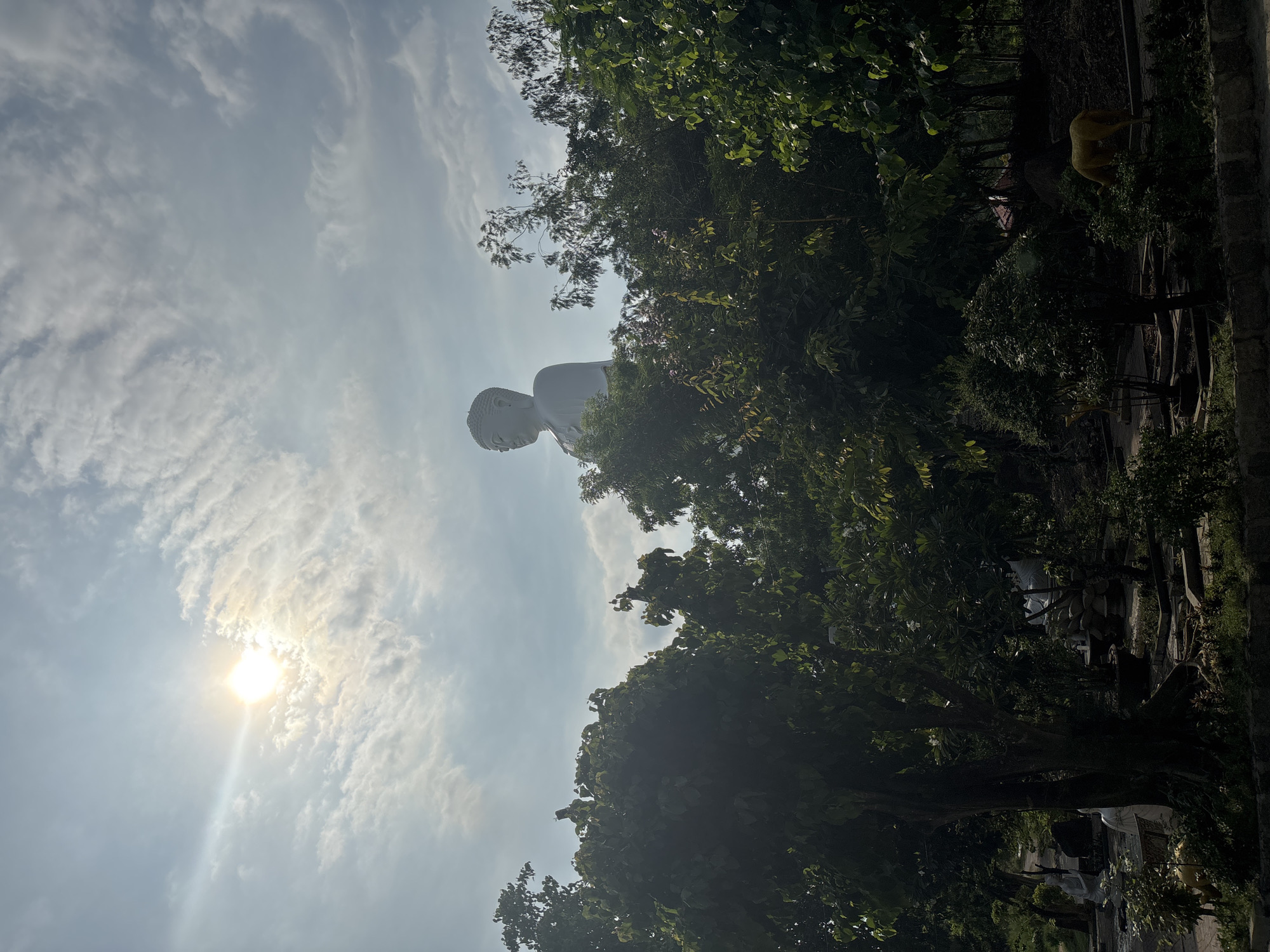 The image depicts a large white statue of a Buddha figure seated in meditation, positioned on a hill and partially obscured by lush green trees. The sun is shining brightly in the sky, partially obscured by wispy clouds, casting a lens flare. In the foreground, there are more trees and vegetation, with some smaller statues and landscaping visible at the base of the hill. A yellow statue of a deer is also present in the lower right portion of the image. The scene appears to be an outdoor religious or park-like setting.