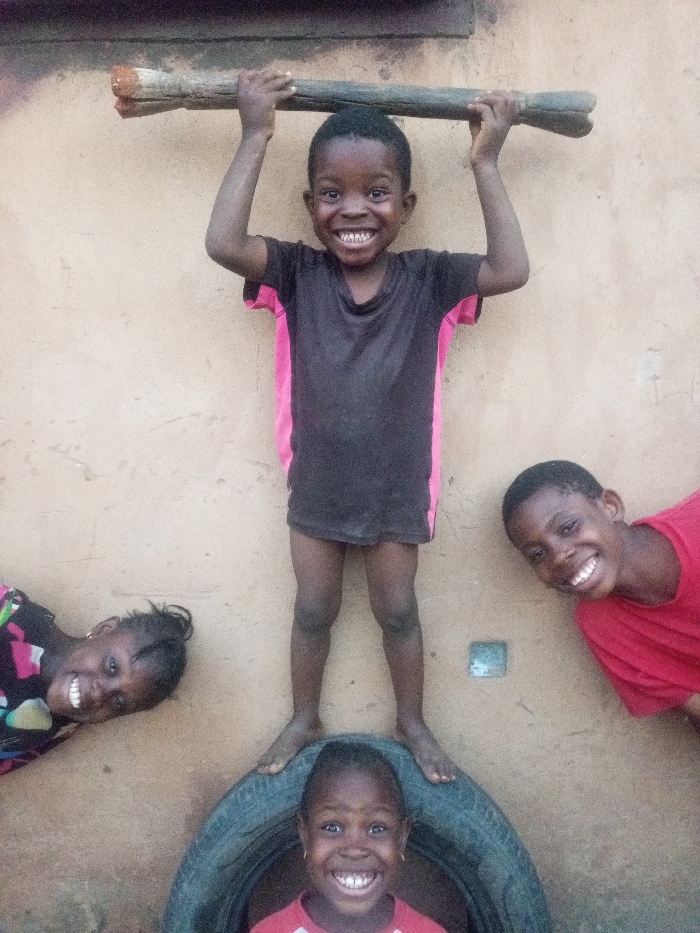 The image captures four young Black children engaged in playful interaction, all displaying wide, joyful smiles against a plain, light brown wall.
In the upper center of the frame, a child with short dark hair stands barefoot, facing the camera. They wear a dark t-shirt with pink panels on the sides and are holding a thick, weathered piece of wood or a branch horizontally above their head with both hands. Their expression is one of pure happiness.
Directly below this child, another young child is positioned inside an old, dark tire, with only their head and upper torso visible. This child also has a bright, open-mouthed smile and appears to be wearing a red and white top. The feet of the child standing above are placed on the rim of the tire.
To the left of the central arrangement, a young girl with braided hair is lying on her side, her head tilted upwards, looking towards the camera with a cheerful smile. She is wearing a colorful top with various patterns, including pink.
On the right side of the frame, another young boy is seen peeking in, his head and shoulder visible. He has short dark hair and is wearing a red t-shirt, also smiling widely towards the camera.
The background consists solely of a simple, unadorned light brown wall, possibly made of mud or concrete, which provides a neutral backdrop, emphasizing the children. The lighting suggests it is daytime. The overall mood of the image is lighthearted and joyful, capturing a moment of genuine happiness and camaraderie among the children. No text is visible in the image.