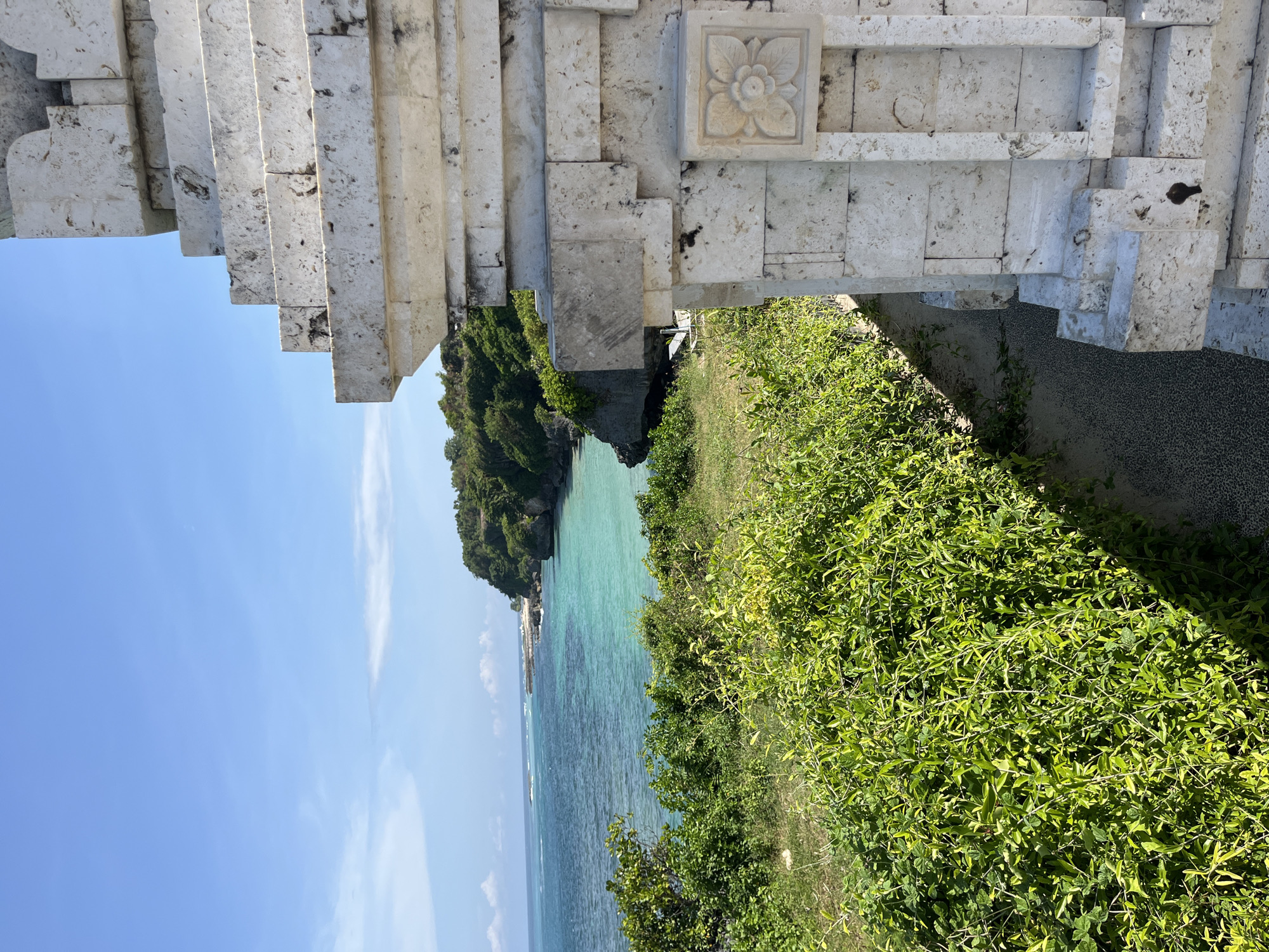 The image captures a coastal view framed by a stone structure, potentially a gate or part of a temple. The location is identified as Kuta Selatan, Indonesia. The stone structure is predominantly white and appears to be made of stacked blocks, with a floral carving on one of the blocks.
Beyond the structure, the view opens to a body of water that transitions in color from turquoise near the shore to a deeper blue further out. A rocky landmass covered in dense green vegetation juts into the water. The sky is clear and blue. The foreground is dominated by green vegetation on what appears to be a hillside. A small patch of gravel is visible between the vegetation and the structure. No people are visible in the image.
