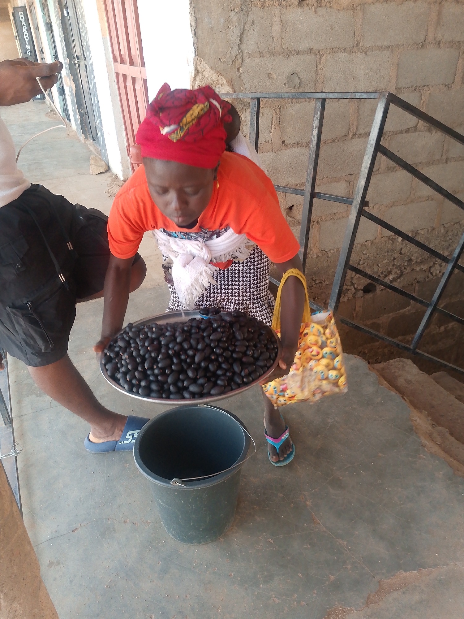 A woman stands holding a round tray piled high with dark, oval fruits, likely olives, over a gray bucket. She is wearing an orange t-shirt, a patterned dress, and a red headscarf. A white cloth is tied around her neck. Beside her, a man wearing a white t-shirt, black cargo shorts, and blue sandals holds the tray. The environment includes a concrete floor, brick walls, a metal railing, and a shopping bag with emoji faces is nearby. The location is Unknown, Unknown.