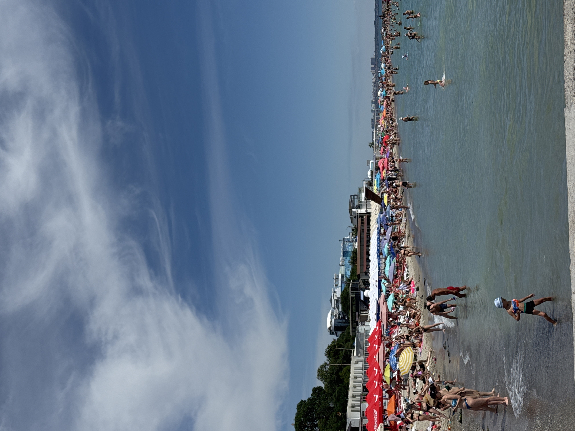 This is a wide-angle, full-shot photograph of a crowded beach scene in Odesa, Ukraine, on a clear, sunny day with scattered wispy clouds in a bright blue sky. The foreground shows the shallow, calm, clear turquoise water with gentle waves lapping at the shore. Several people are in the water, including a child in the lower right foreground wearing a hat and holding a toy gun. Further out in the water, several adults are swimming and standing.
The middle ground is a densely populated beach lined with rows of colorful beach umbrellas, predominantly red, white, and blue, providing shade for beachgoers. People are seen sunbathing on towels and lounge chairs, walking along the shore, and interacting with each other. The sand appears light in color.
In the background, there are various buildings and structures along the coastline. These include some lower-rise buildings with white facades, possibly beachside cafes or changing rooms, and further back, taller, modern buildings that form part of the Odesa skyline. Trees are visible on the left side of the image, suggesting vegetation beyond the immediate beach area. The overall impression is a bustling summer scene at a popular seaside resort.
