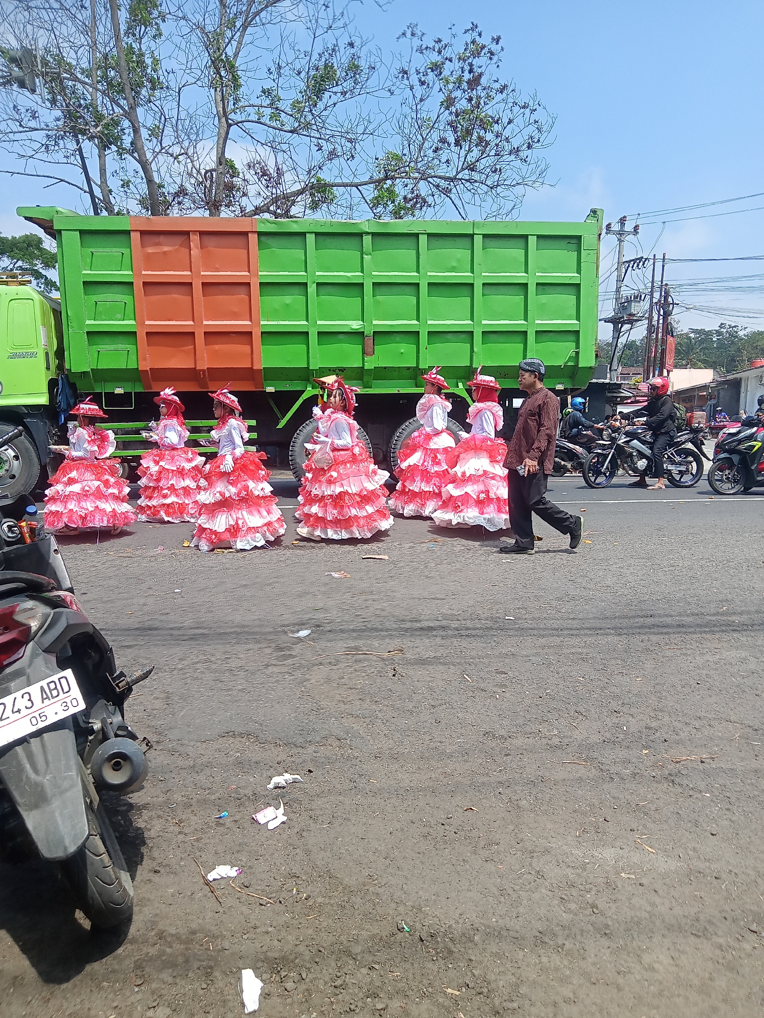 On a sunny day in Urutsewu, Indonesia, a line of performers in ornate white and red ruffled dresses and matching hats stands in front of a large green dump truck with an orange cargo section. To the right of the performers, a man in a brown and white striped shirt and dark pants walks away from the truck, holding a dark object in his right hand. In the background, several motorcycles are parked on the side of the road, with riders wearing helmets. The scene unfolds on a paved road with scattered debris. A parked motorcycle is in the foreground on the left side of the image.