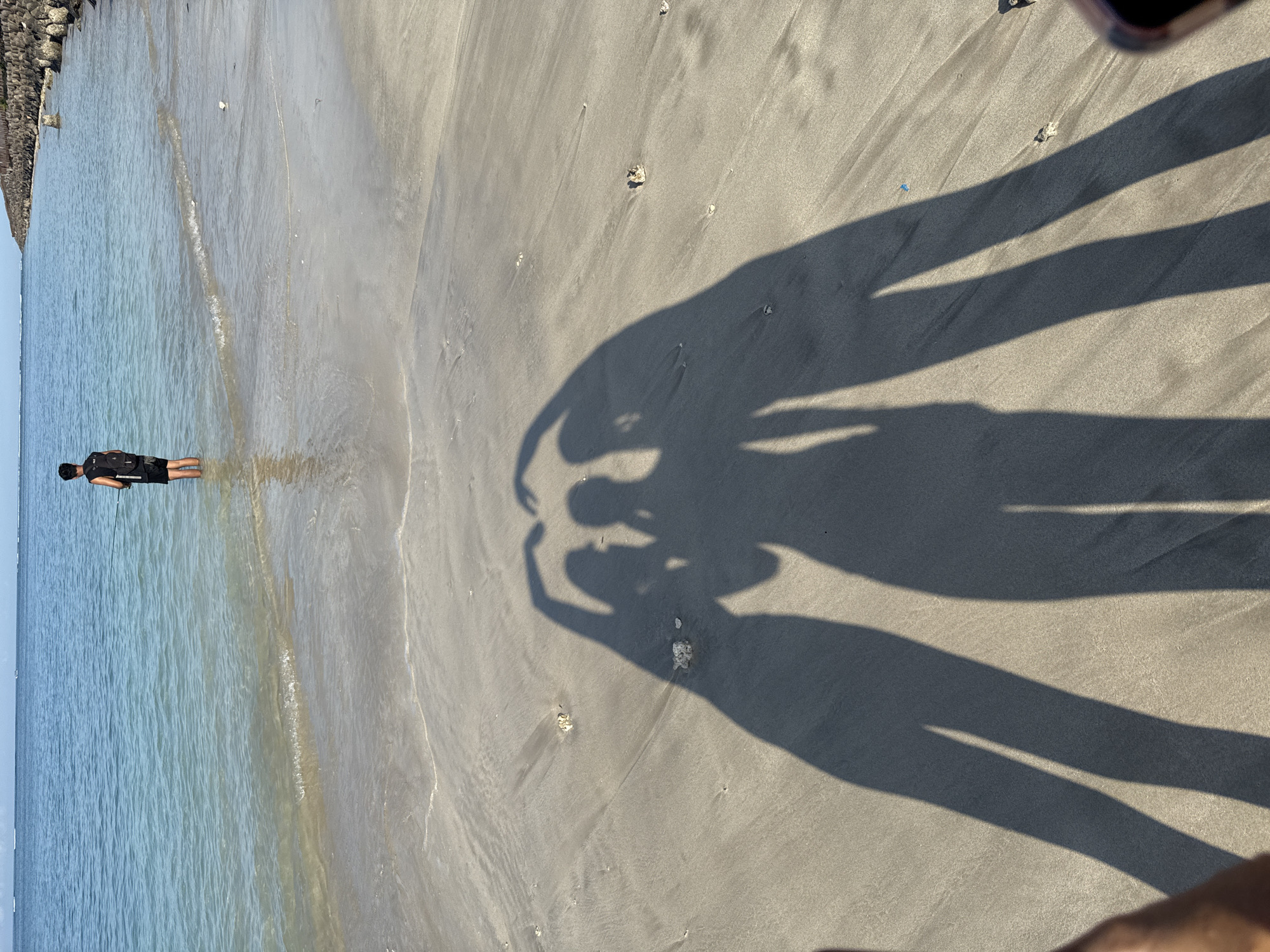 The image shows a beach scene with a person standing in the water along the shoreline on the left side of the image. The water is blue-green and appears shallow near the shore. To the left is a rock wall. On the sandy beach, there are shadows cast by two individuals, appearing to form a heart shape with their hands overhead. The beach is light gray-brown and has scattered shells. The image may be taken in Kuta, Indonesia.