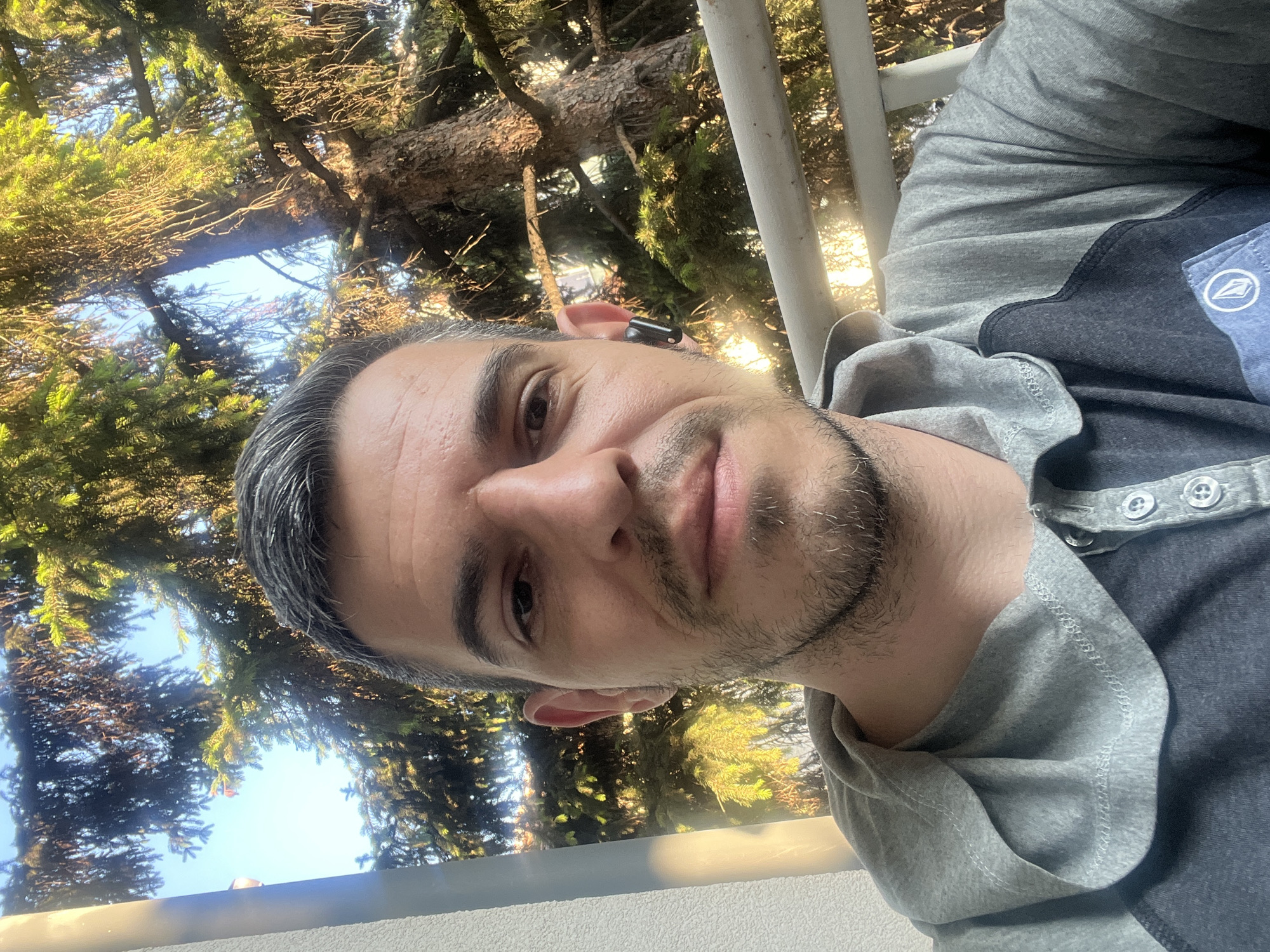 A man with short, graying hair and a goatee is taking a selfie outdoors. He is wearing a gray hoodie with dark blue accents and a white logo patch on the chest. He has a black wireless earbud in his right ear. Behind him, a large evergreen tree is visible, with the sky and some buildings in the background. The lighting suggests it is daytime, possibly late afternoon given the warm glow. The scene appears to be from a balcony or outdoor seating area. This setting is consistent with a residential area in Sofia, Bulgaria, where such balconies and mature trees are common.