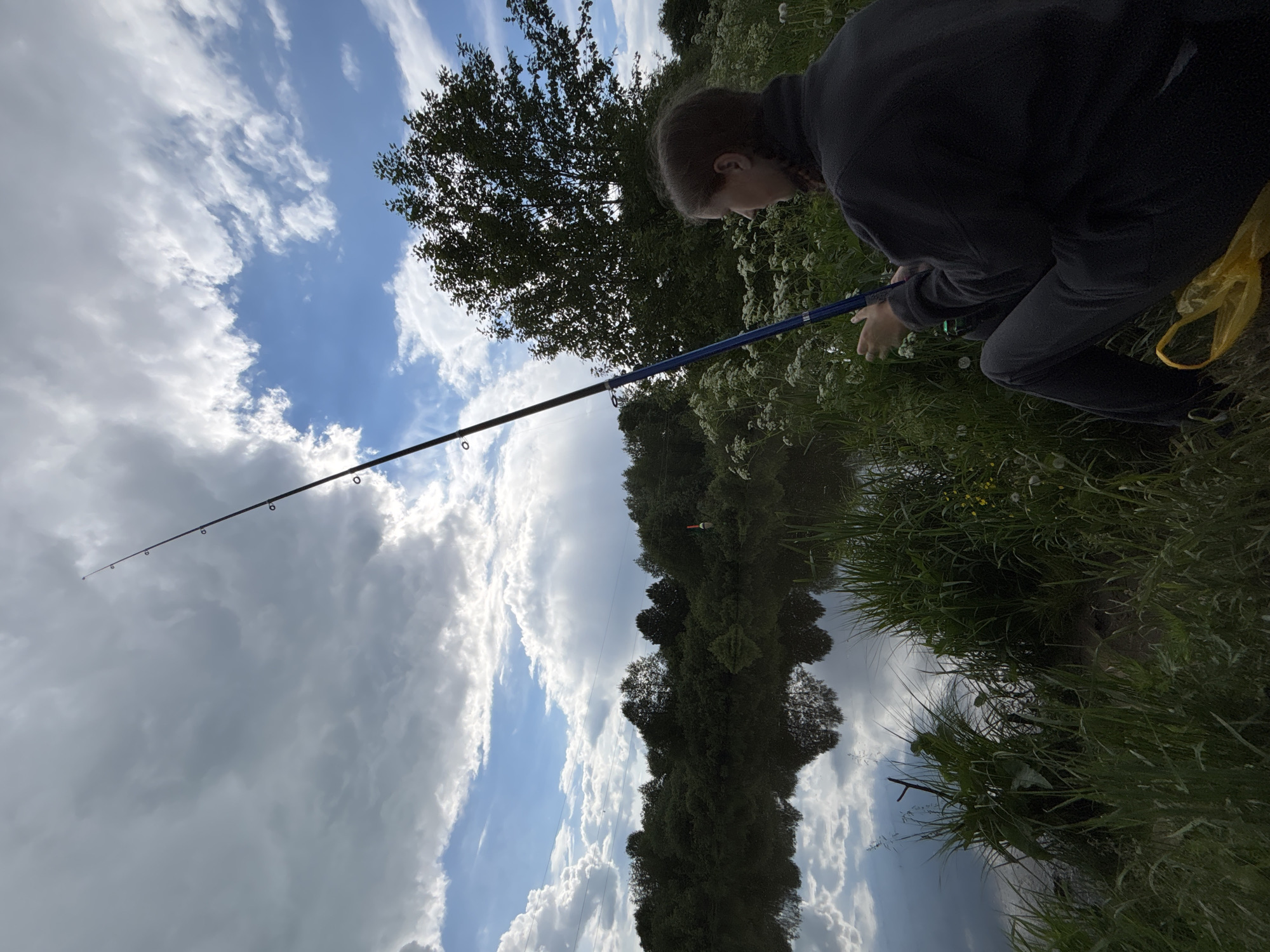 A young individual is fishing along a grassy bank next to a body of water reflecting the sky. The person is wearing dark clothing and is holding a blue fishing rod. A yellow bag is near them. The sky is visible with both clear blue patches and large cloud formations. Trees are present, both along the bank and reflected in the water. The image appears to be taken outdoors in a natural setting. Фоки, Russia, is not discernible from the visual details.