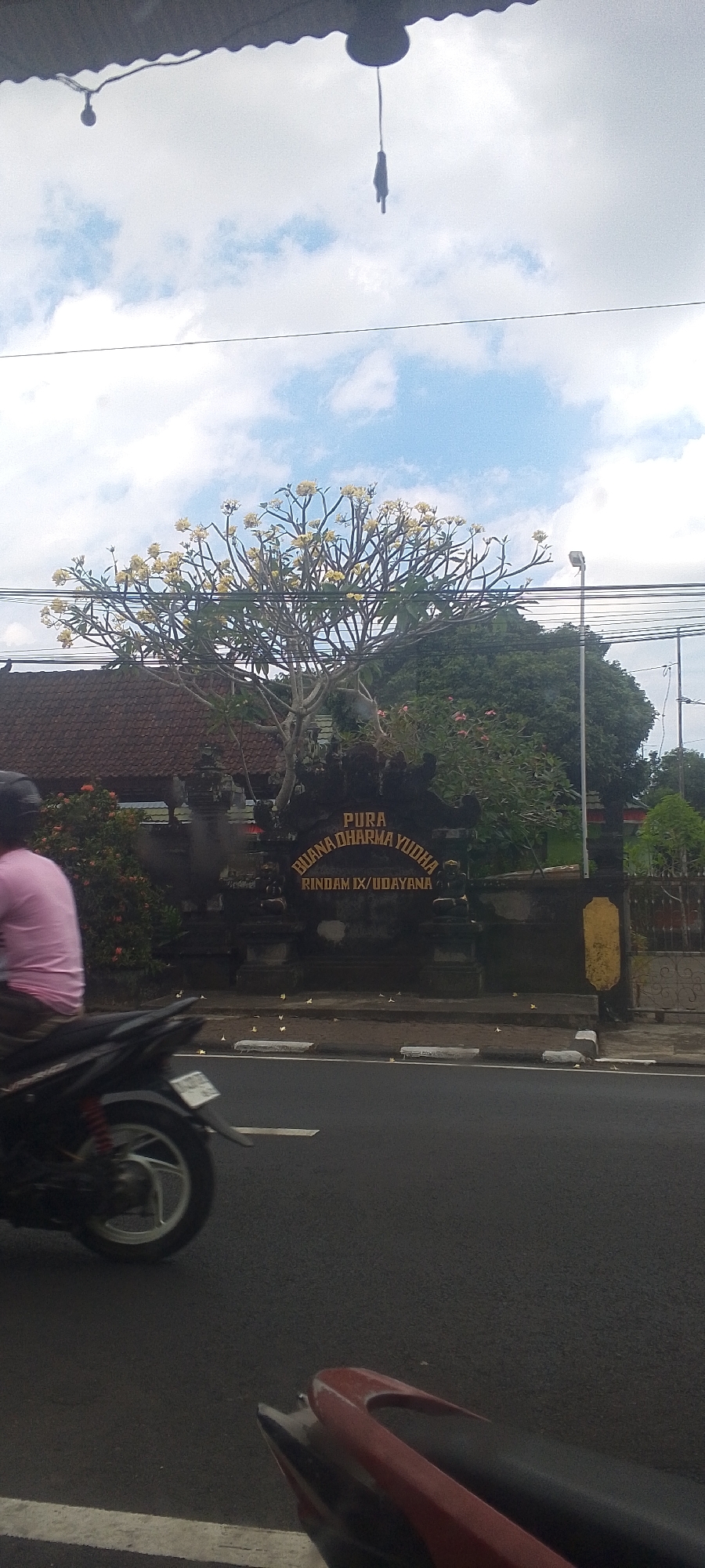 In Kediri, Indonesia, a street scene unfolds under a partly cloudy sky. A motorcycle, partially visible in the foreground with its rear wheel on the left, is on a dark asphalt road marked with white lines. A person wearing a pink shirt is seated on a black motorcycle, also partially visible on the left side of the frame, moving away from the viewer. Behind the road, a dark stone gateway with gold lettering that reads "PURA BUANA DHARMA YUDHA RINDAM IX/UDAYANA" stands prominently. To the left of the gateway, a traditional Balinese-style house with a red tiled roof is visible. In front of the gateway, a tree with delicate yellow flowers is in bloom, its branches reaching towards the sky. The environment appears to be a public street with overhead electrical wires and a lamppost visible in the background.