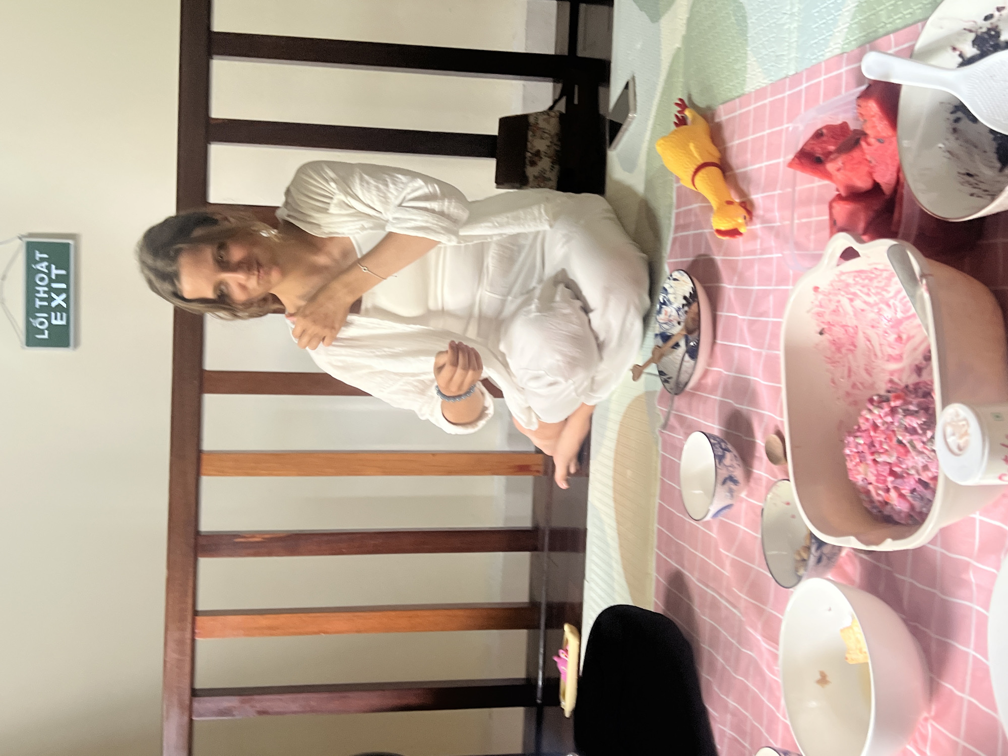 A woman in white clothing sits cross-legged on a floor covered by a pink and white checkered mat. Behind her, a wooden railing separates the space from a wall with a green exit sign. In front of her, various bowls of food are arranged, including a large bowl of pink salad, a smaller bowl with what appears to be chickpeas, and a bowl containing cubed watermelon. A yellow rubber chicken toy lies on the mat near the watermelon. A smartphone is visible on the floor to the right. The scene suggests a casual meal or gathering. The presence of the Vietnamese "EXIT" sign indicates the location is likely in Đà Nẵng, Vietnam.