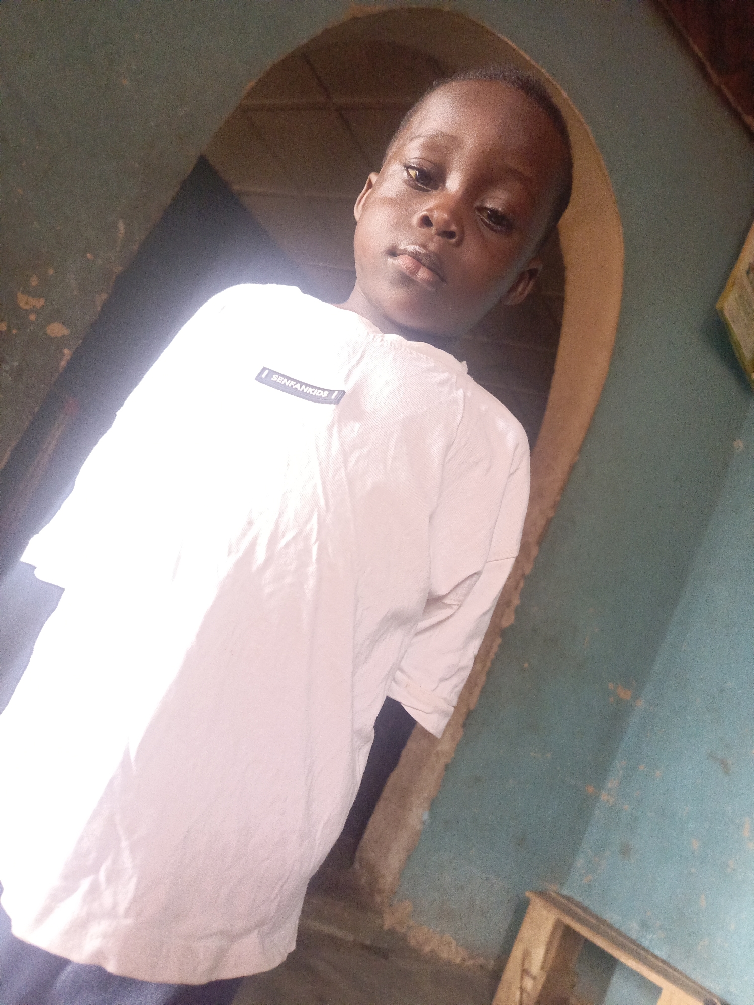 A young boy stands indoors near an arched doorway. He wears a white t-shirt with a "SENFAN KIDS" label. The wall surrounding the doorway is teal. A small, wooden stool sits in the lower right corner of the image. The location is Unknown, Unknown.