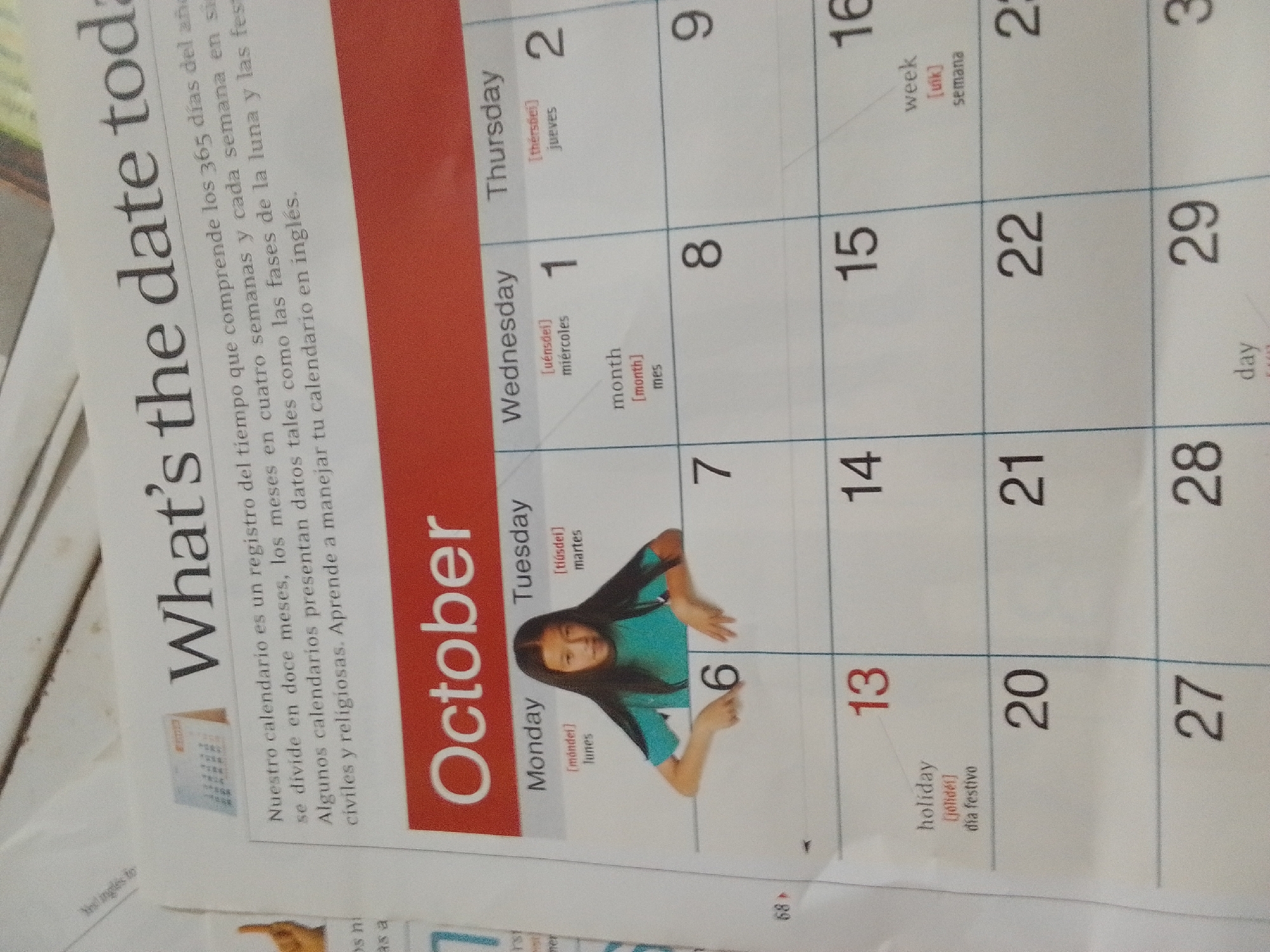 This is a photograph of an open calendar page, part of a publication with the title "What's the date toda". The calendar is for October and features a portion of the months of September and November as well. The calendar is presented in English, with Spanish translations provided for days of the week, months, and terms like "week", "month", "day", and "holiday". The page is oriented such that the text is mostly readable.
The prominent subjects are the calendar grid itself, and a young girl with long dark hair, wearing a bright teal shirt, is partially obscuring the grid from the 6th to the 13th of October. She is looking at the camera with a neutral expression, and her right hand is extended, touching the grid near the number 6. The calendar design is clean and functional, with the month "October" highlighted in a red block on the left side.
The text on the page includes the title "What's the date toda", and a paragraph in Spanish explaining that the calendar is a record of time, divided into twelve months, four weeks per month, and covering lunar phases and civil/religious holidays, with instructions to learn to manage your calendar in English. The days of the week are labeled from Monday to Sunday, with some numbers of the calendar visible: 1, 2, 6, 7, 8, 9, 13, 14, 15, 16, 20, 21, 22, 27, 28, 29. The number 68 is visible in small print at the bottom right of the page.
The scene appears to be an educational or informational context, possibly from a textbook or a learning resource. The lighting is bright, suggesting it might be daytime, and the setting is indoors given the page and calendar format. There are no strong indicators of weather or a specific outdoor location. The image is cropped to focus on the calendar and the girl. The location context "La Tagua, Venezuela" is provided but not visually evident within the image itself.