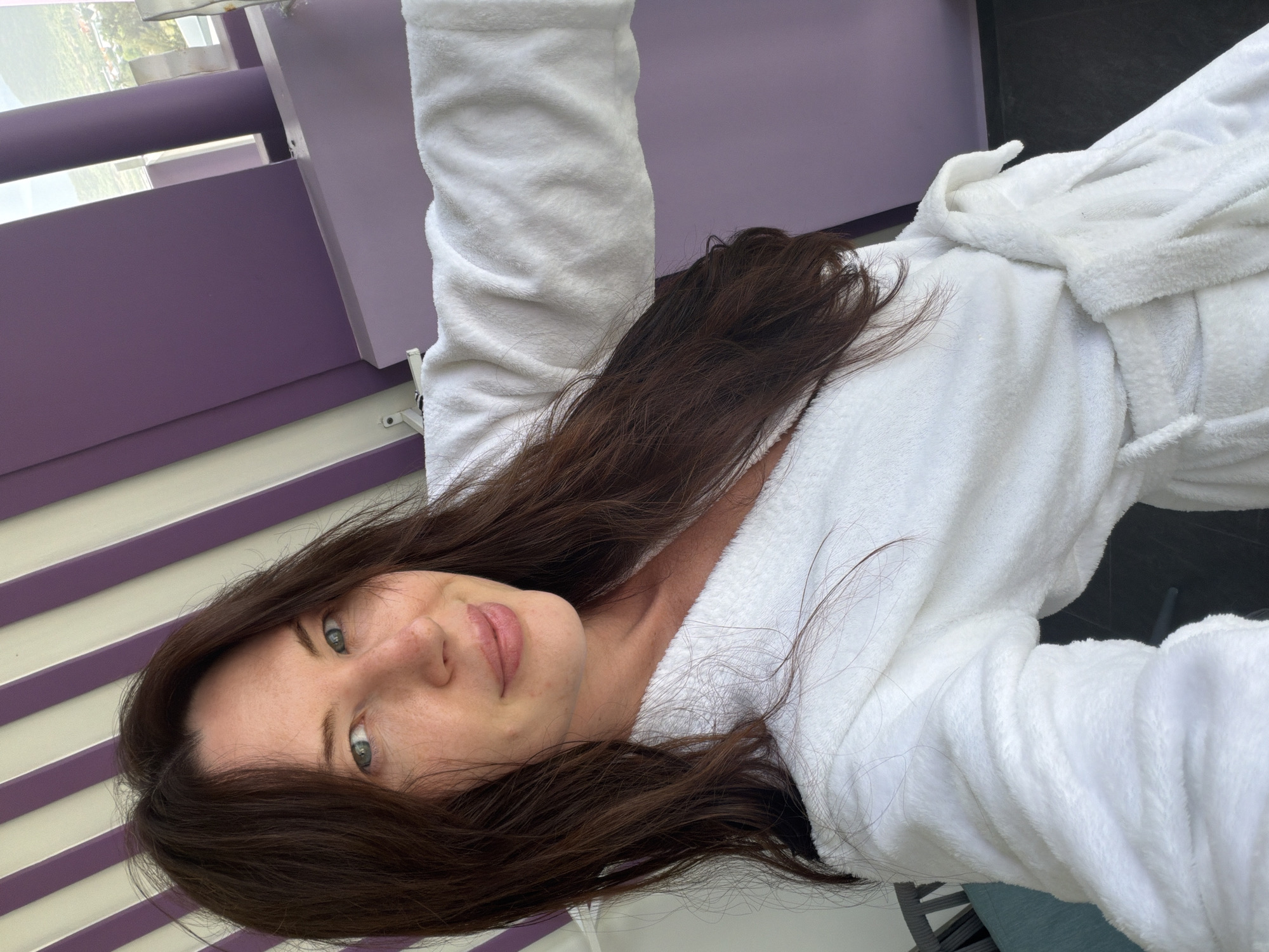 A woman with long, dark brown hair is taking a selfie. She is wearing a white terrycloth robe and appears to be indoors, possibly in a hotel room, with a view of a city or town in the background. The room has distinctive purple and white striped decor. The woman has fair skin, blue eyes, and a light smile. The scene is well-lit, suggesting it is daytime. The image does not provide any information that would indicate it is located in Dong Ninh Hoa Ward, Vietnam.