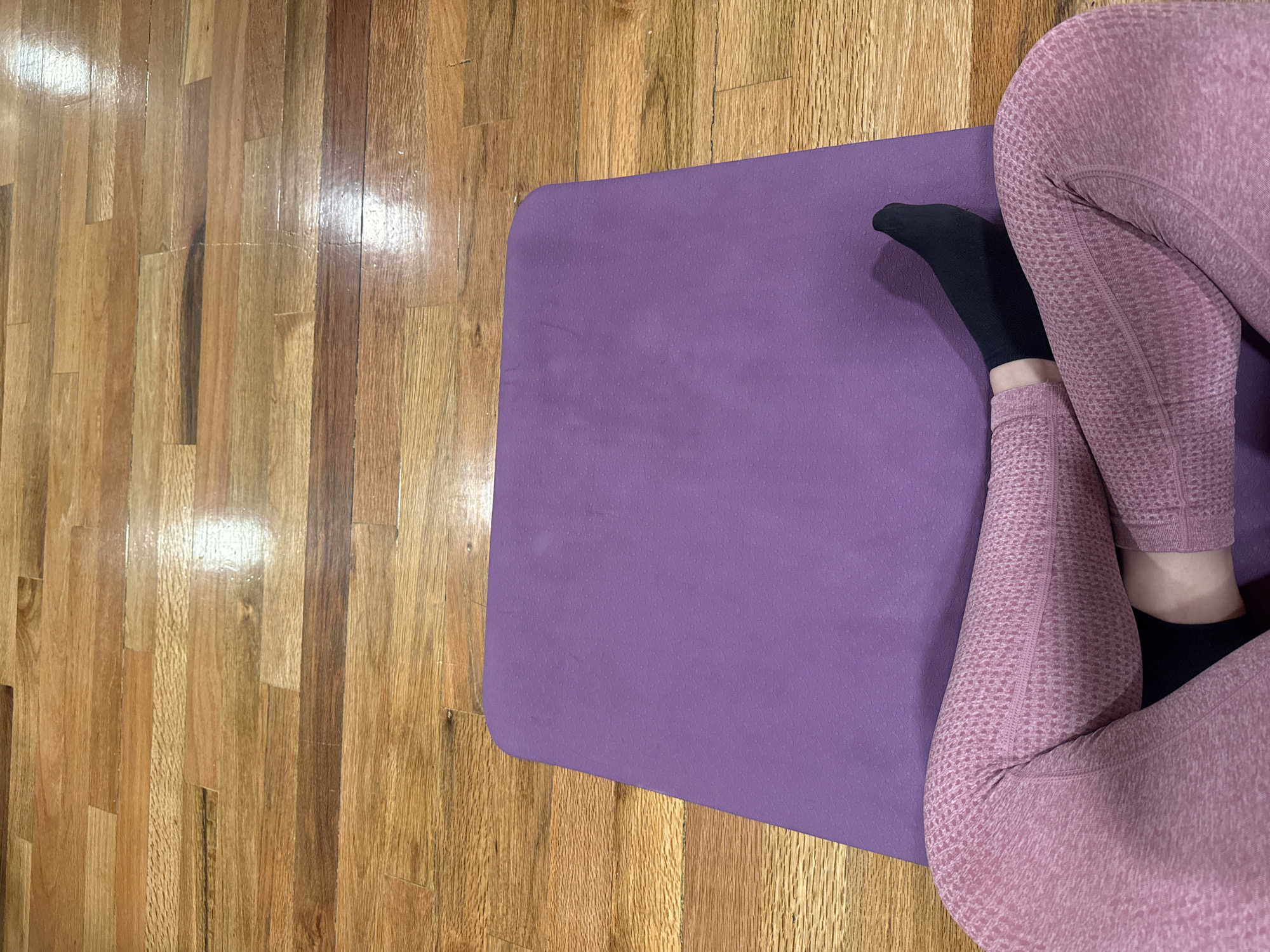 A high-angle shot depicts a person sitting on a purple yoga mat placed on a wooden floor. The individual is wearing pink leggings and black socks. The wooden floor appears to be polished and reflects light. The image does not provide sufficient information to determine the specific location in Puebla, Mexico.