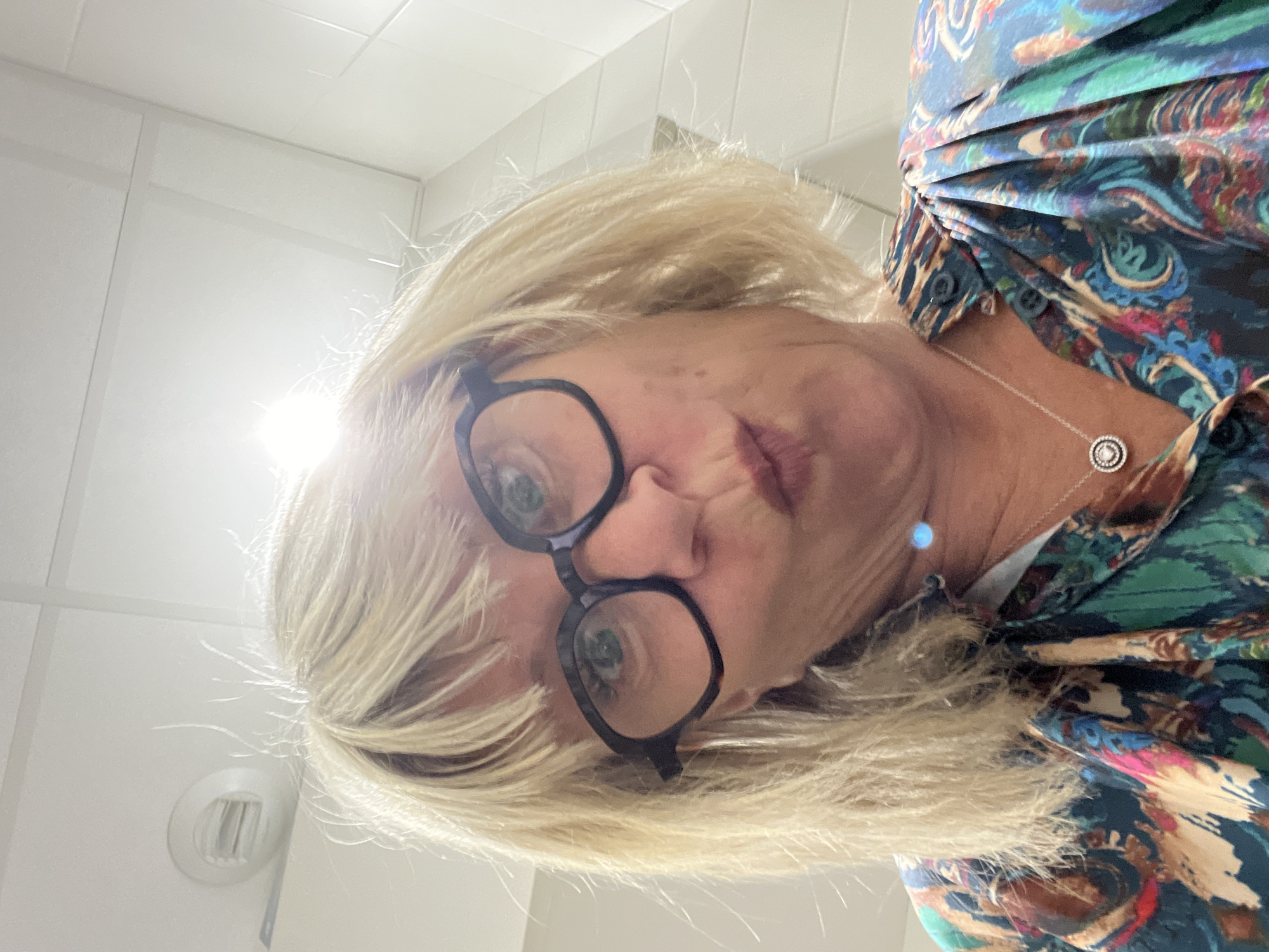A close-up, low-angle shot captures a person with blonde, shoulder-length hair and black-rimmed glasses looking directly into the camera. The individual's head is tilted, and their lips are slightly pursed, conveying a neutral expression. Their eyes are magnified by the glasses, reflecting a hint of green.
The person is wearing a brightly patterned shirt with an intricate design featuring various shades of blue, green, and hints of red, orange, and brown. A delicate silver necklace with a circular, possibly jeweled, pendant is visible around their neck.
The background is simple and bright, consisting of white walls and ceiling tiles, suggesting an indoor setting. A strong light source in the upper left creates a noticeable glare, illuminating the scene. A white circular ventilation grille is visible on the ceiling or upper wall towards the lower left of the frame. The perspective is from below the subject, as if the person is looking down at the camera. No text is visible in the image. The location is La Fouillouse, France.