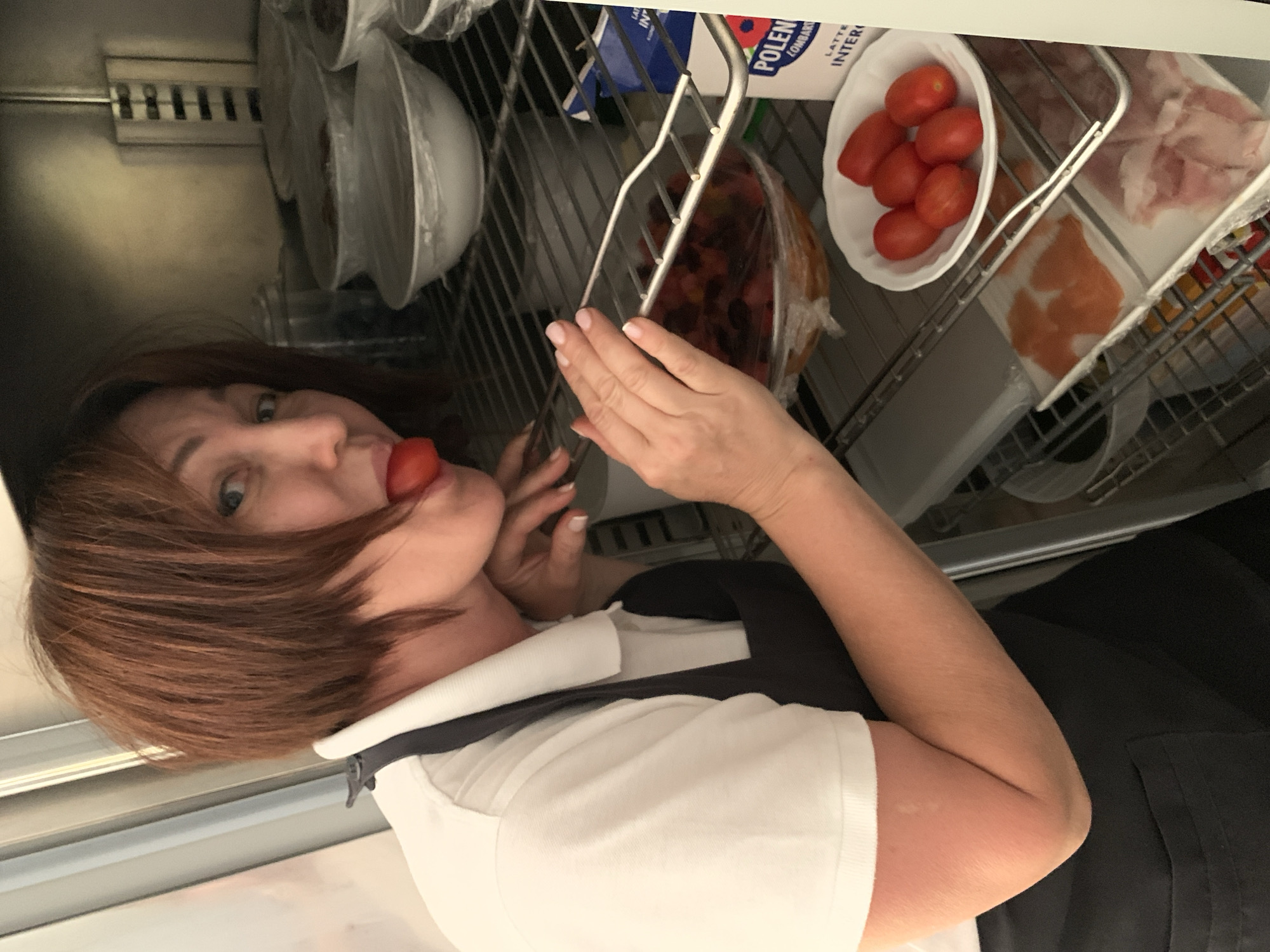 A woman with short, brown hair and a white polo shirt with a black apron is leaning into a commercial-style refrigerator. She is holding a cherry tomato to her tongue and looking towards the camera. The refrigerator shelves contain various food items, including bowls covered in plastic wrap, a bowl of chopped tomatoes, and a plate of cherry tomatoes. Below, there are packages of raw fish and meat. A blue and white carton with Italian text is visible on one shelf. The refrigerator appears to be in a professional kitchen setting. The location is specified as Castiglione d'Orcia, Italy.