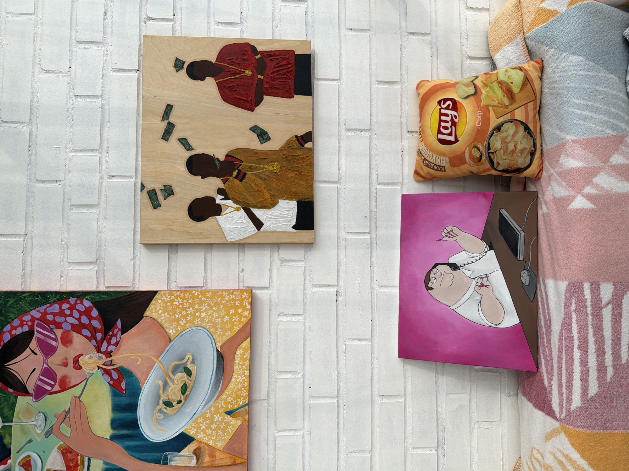 The image depicts a wall adorned with three framed artworks and a decorative pillow, set against a white brick wall. The uppermost artwork on the left is a painting of a woman with dark hair, wearing a red and purple polka dot headscarf and pink sunglasses, eating spaghetti from a white bowl. She is dressed in a yellow floral print dress. To the right of this painting is a wooden board with a painting of three dark-silhouetted figures, two wearing gold chains, with green dollar bills scattered around them. The bottom left artwork is a painting of a cartoon character, resembling Peter Griffin, sitting at a desk and talking on the phone. Beside this painting, on the right, is a pillow shaped like a bag of Lay's potato chips. The artworks and pillow are placed on top of a knitted blanket draped over a surface, possibly a sofa or bench. The lighting suggests an indoor setting, likely a home or gallery. There are no visible signs or indicators that the location is in Krasnoyarsk, Russia.