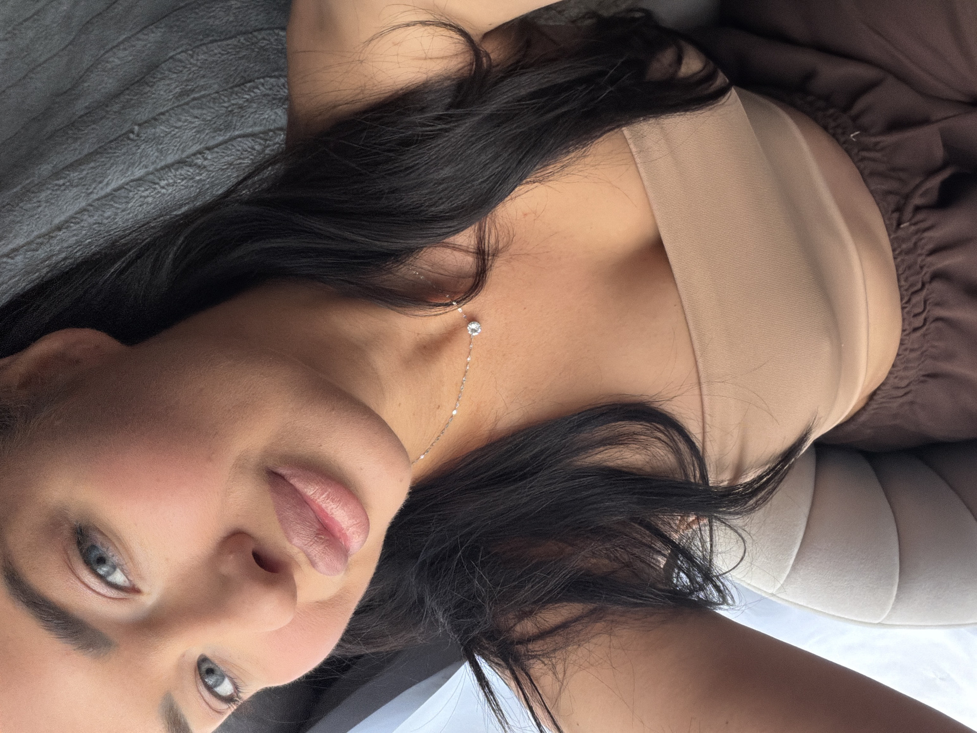 A woman with long, dark hair and light eyes is lying on a grey, textured surface next to a cream-colored cushion and brown fabric. She is wearing a beige top and a delicate necklace with a small, sparkling pendant. Her face is directed towards the camera, displaying soft makeup with emphasis on her lips. The image does not contain information related to the location being in the city of Moscow, Russia.
