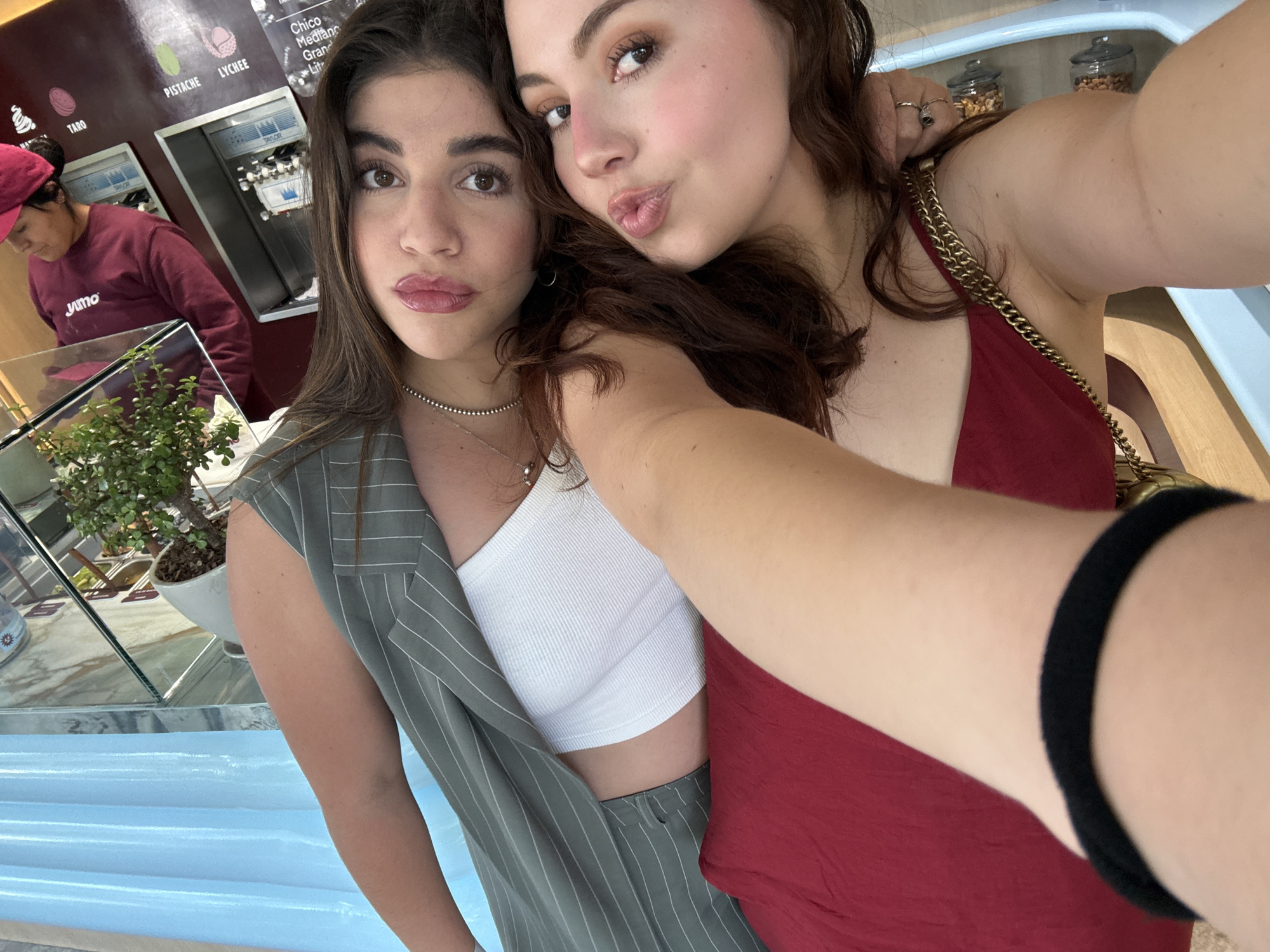 Two young women are posing closely for a selfie indoors, likely in an ice cream shop in Mexico City, Mexico. The woman on the left has dark hair, light skin, and is wearing a white crop top with a gray pinstriped vest. She has a silver necklace and is looking at the camera with a slight pout. The woman on the right has reddish-brown hair, fair skin, and is wearing a red tank top. She is leaning towards the camera with her lips puckered in a kiss. The camera is close to her face.
In the background, an employee wearing a red shirt and pink hat stands behind a counter. A sign in the background lists flavors like Pistache and Lychee. Ice cream machines are also visible. A glass display case with a small potted plant is in the foreground. Jars filled with various toppings are visible on a shelf behind the woman in red.