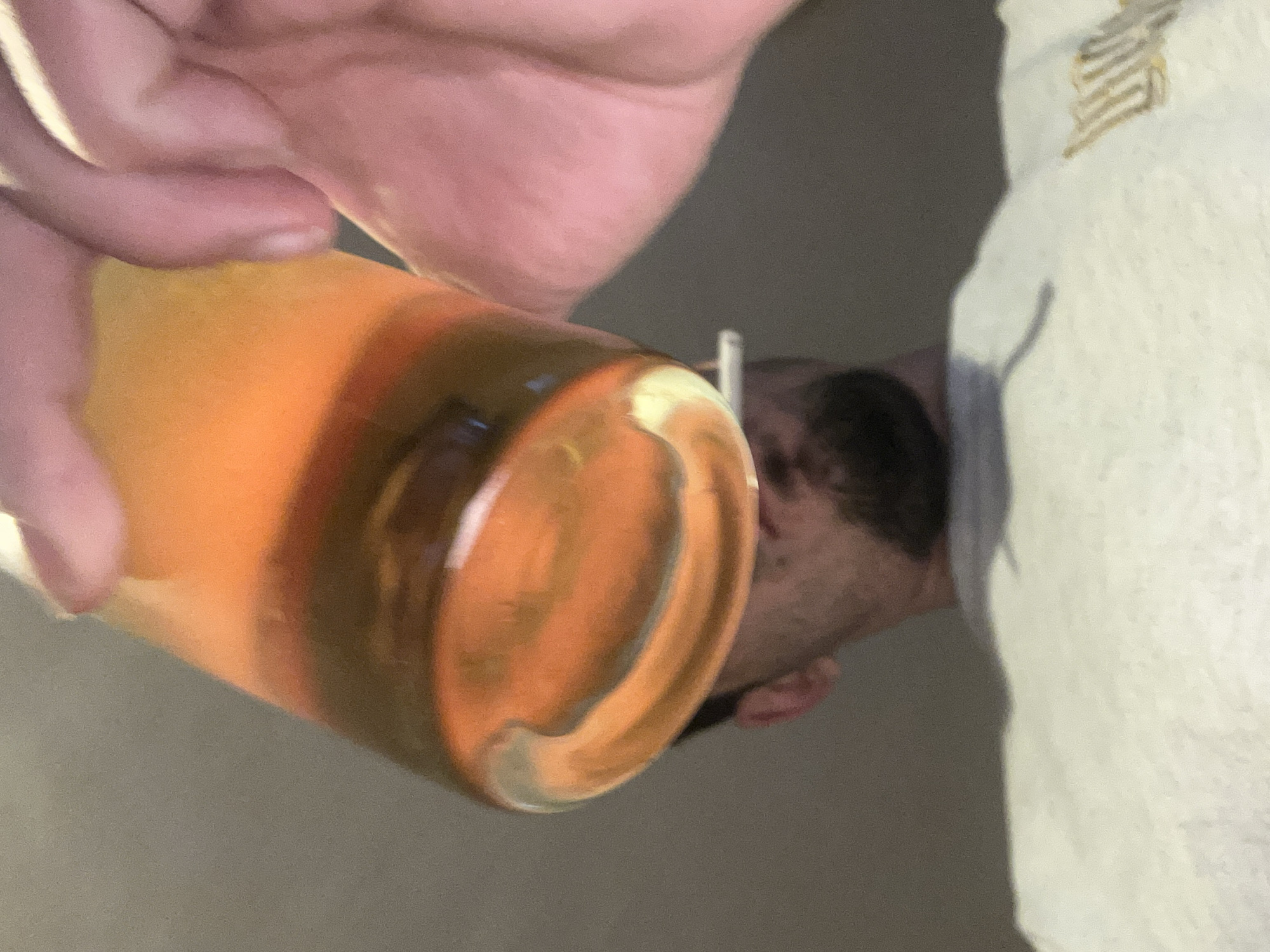 The image, rotated onto its side, captures a close-up perspective of a person's hand holding a glass. The hand, with light skin, is gripping the upper part of a clear, cylindrical glass.
The glass contains a layered beverage, appearing bright orange and yellow in its upper section and transitioning to a darker, caramel-brown color towards the bottom. The glass surface reflects light, showing some highlights.
In the blurred background, the profile of another person's head is visible. This individual has dark hair and a light complexion, with part of their ear and a faint outline of their face discernible. To the right of the frame, a light-colored, textured fabric, possibly white or off-white, is present, displaying an intricate, golden-brown embroidered design.
The activity depicted is the holding of the glass, suggesting an imminent or recent act of drinking. The scene is illuminated by soft, warm light, creating shadows that add depth, consistent with an indoor setting. No discernible text is visible within the image. The image is contextualized to Varna, Bulgaria.