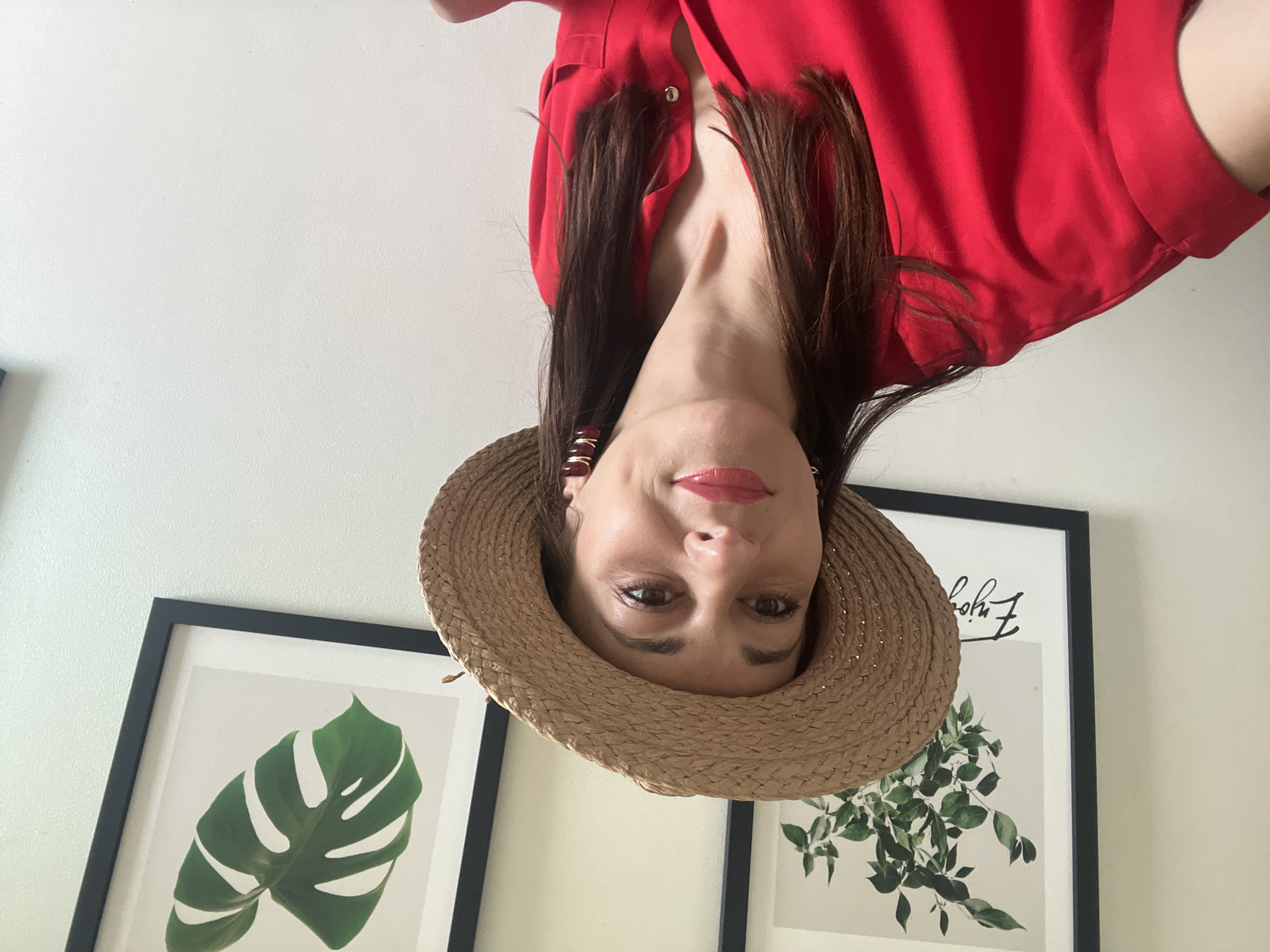 A woman is taking a selfie in a room with two framed botanical prints on the wall. She is wearing a red shirt and a straw hat. The woman has dark hair and is wearing large earrings. The prints are of a plant with leaves and a monstera leaf. The wall appears to be white.