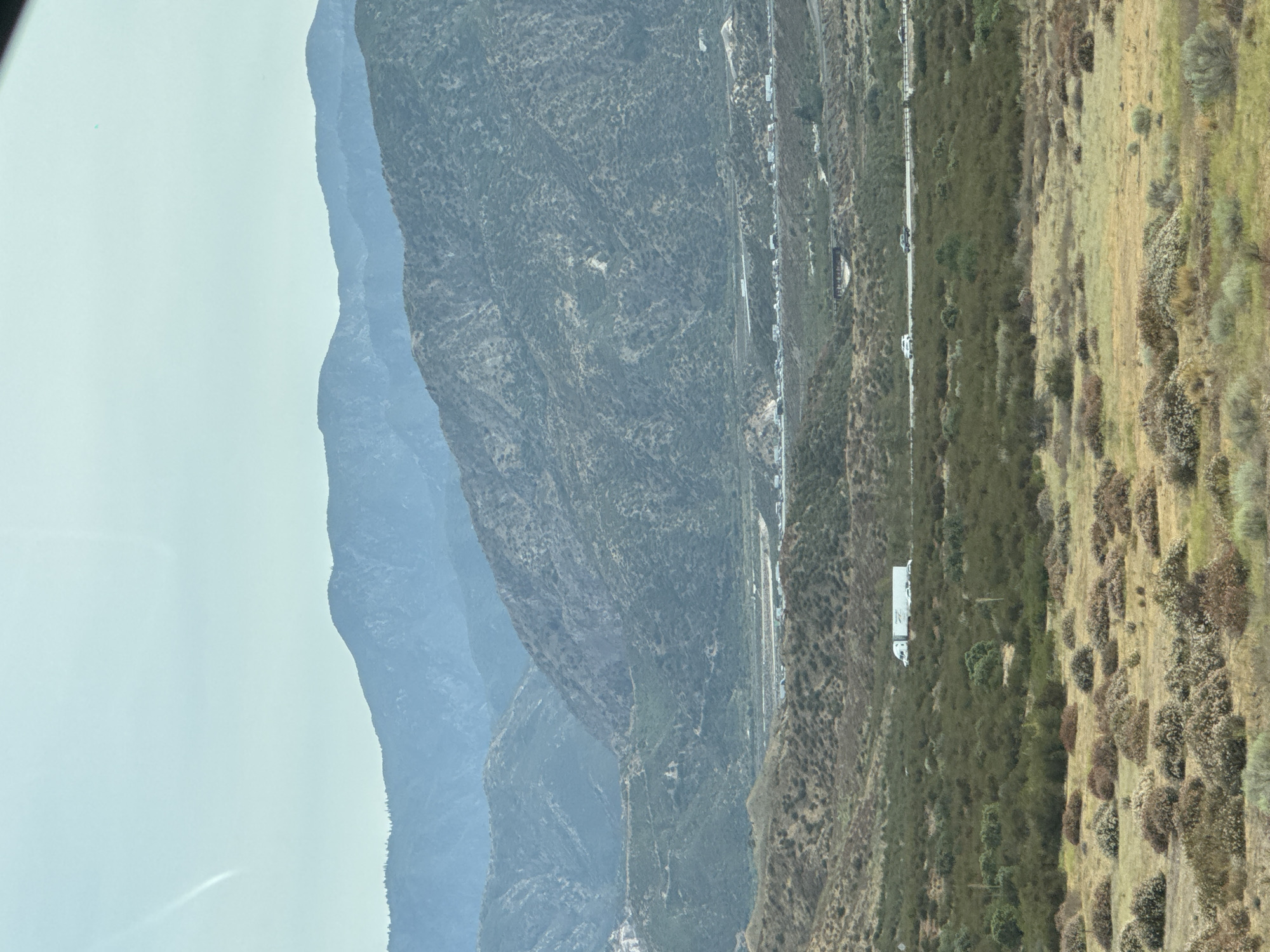 The image depicts a wide, panoramic view of the city of Oak Hills, California, United States, characterized by rolling hills and a hazy sky. In the foreground, dry, golden grass dotted with scattered shrubs and bushes covers the terrain. As the scene recedes, the landscape transitions to more densely vegetated, green hills. Further in the distance, a highway is visible, with several vehicles, including a white truck and cars, traveling along it. Beyond the highway, a congestion of vehicles indicates potential traffic. The background is dominated by layered mountain ranges that fade into a soft blue hue due to atmospheric haze, with the sky above being a pale, light blue.