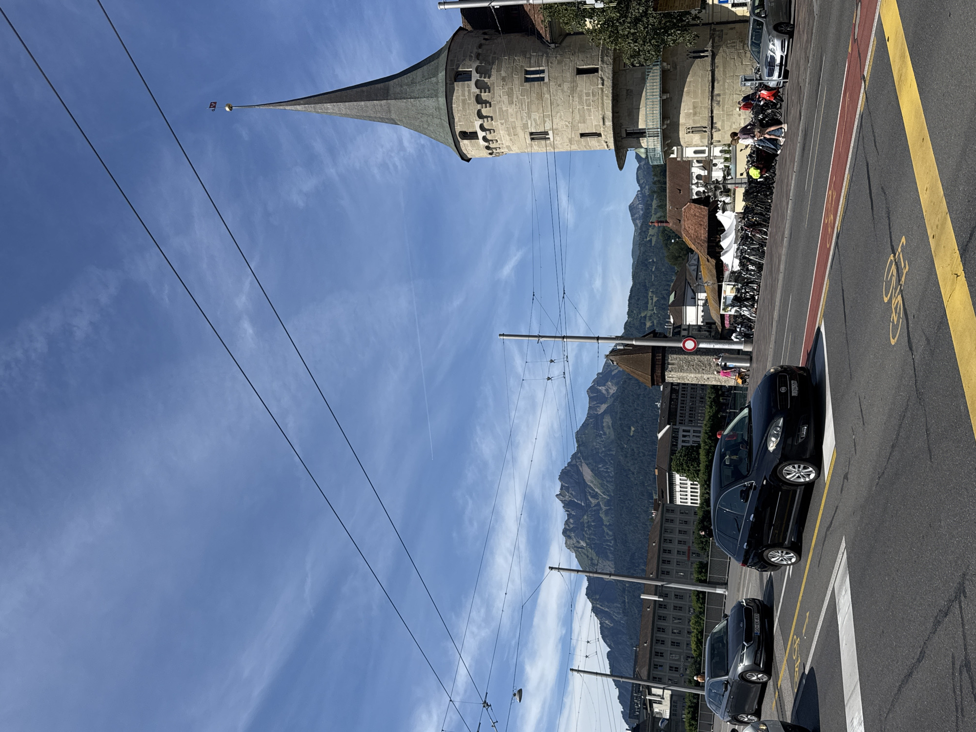 The image captures a street scene in Lucerne, Switzerland, with the iconic Chapel Bridge (Kapellbrücke) and Water Tower (Wasserturm) visible in the background. A cobblestone street with bicycle lanes is in the foreground, with a few cars driving on it. A black Fiat 500L station wagon is in the right lane, and a gray Audi A4 is in the left lane. Several people are walking on the sidewalk on the right side of the street, near a row of parked bicycles. In the background, the Chapel Bridge spans the Reuss River, and the Water Tower stands tall next to it. The mountains of central Switzerland are visible in the distance, with a clear blue sky above.