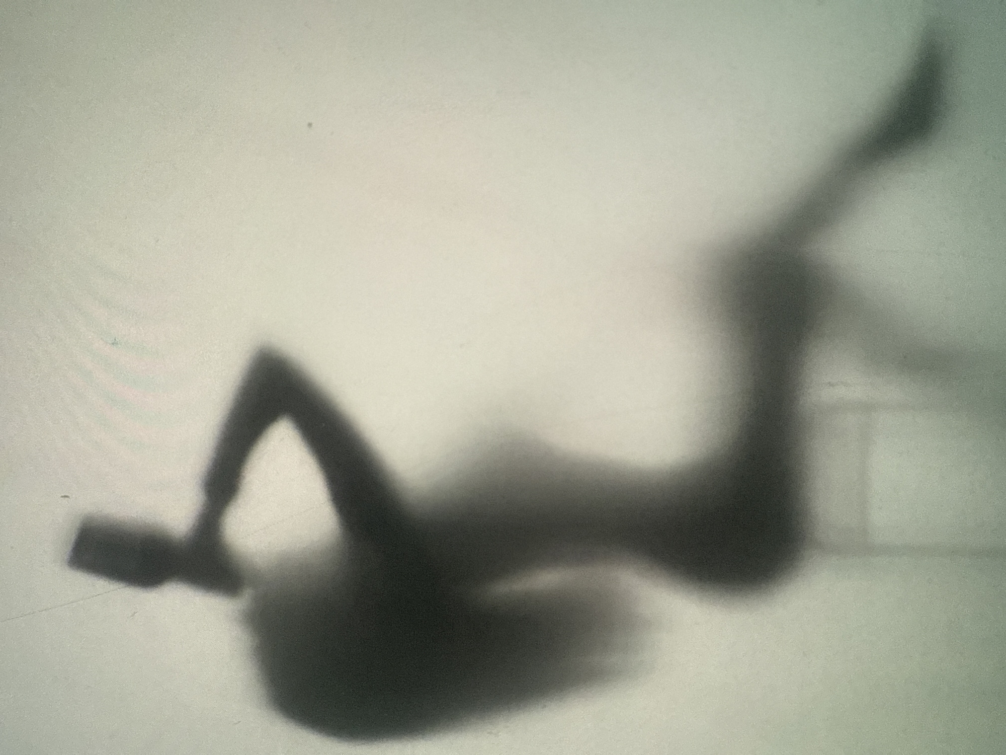 A silhouette of a person is visible through a translucent barrier, possibly frosted glass or a sheer curtain. The person appears to be sitting on a chair, with their body angled to the left. Their left arm is raised, with their hand near their head, and their right arm is bent, resting on their lap. Long hair flows down their back. The background is a light, neutral color with a subtle textured appearance. The lighting is soft, creating a diffused and somewhat dreamlike quality. The scene is abstract and lacks specific environmental or city details.