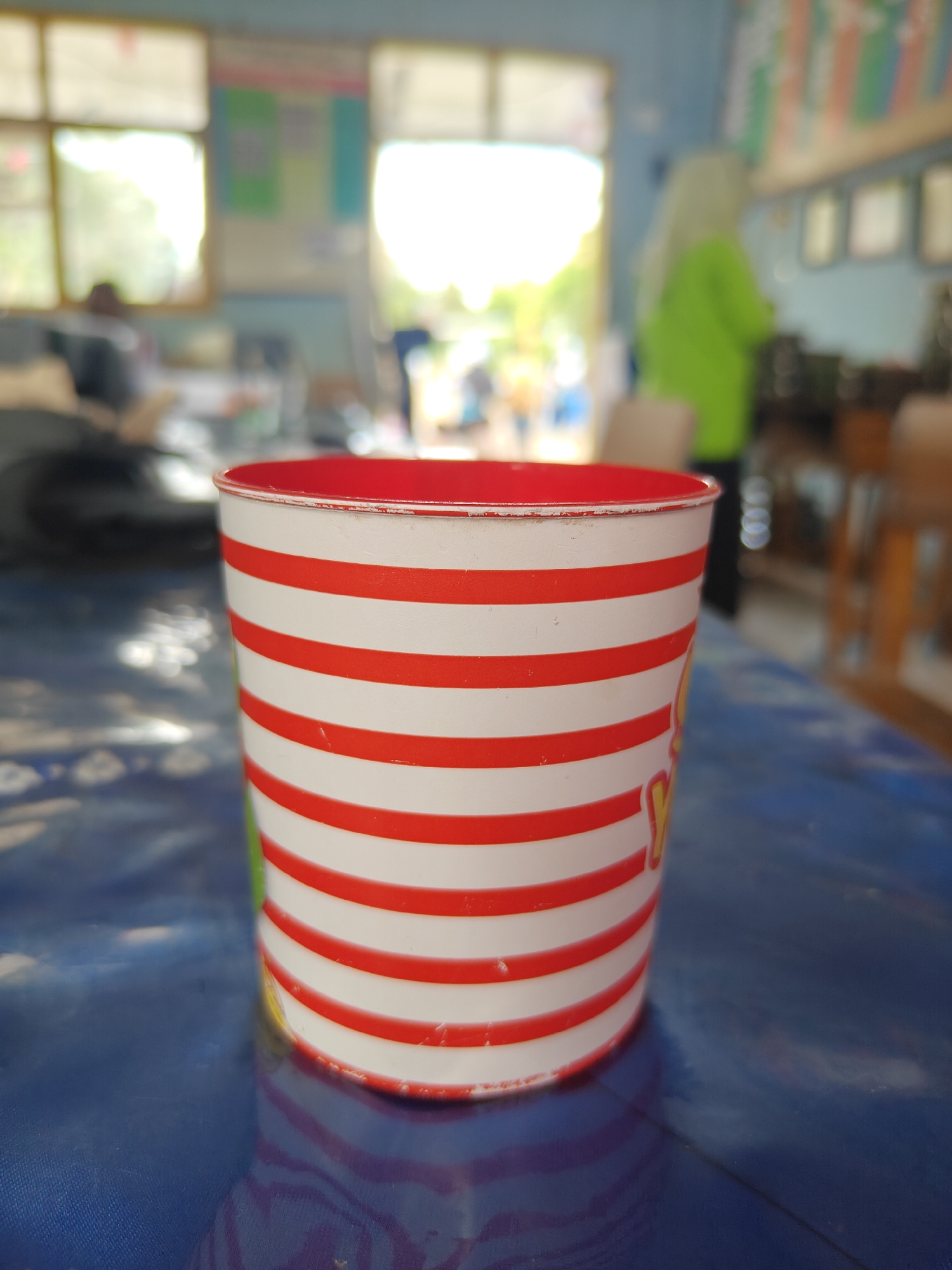 A striped paper cup with red and white horizontal stripes sits on a blue textured surface. The cup is positioned in the foreground, slightly off-center to the left. The background is blurred, suggesting an indoor environment, possibly a classroom or a community hall in Cermee, Indonesia. In the background, figures can be seen, including one person wearing a green garment and a headscarf, and others seated at tables or walking. Sunlight streams in from windows, creating bright areas and casting shadows. Educational posters or charts are visible on a blue wall in the background.