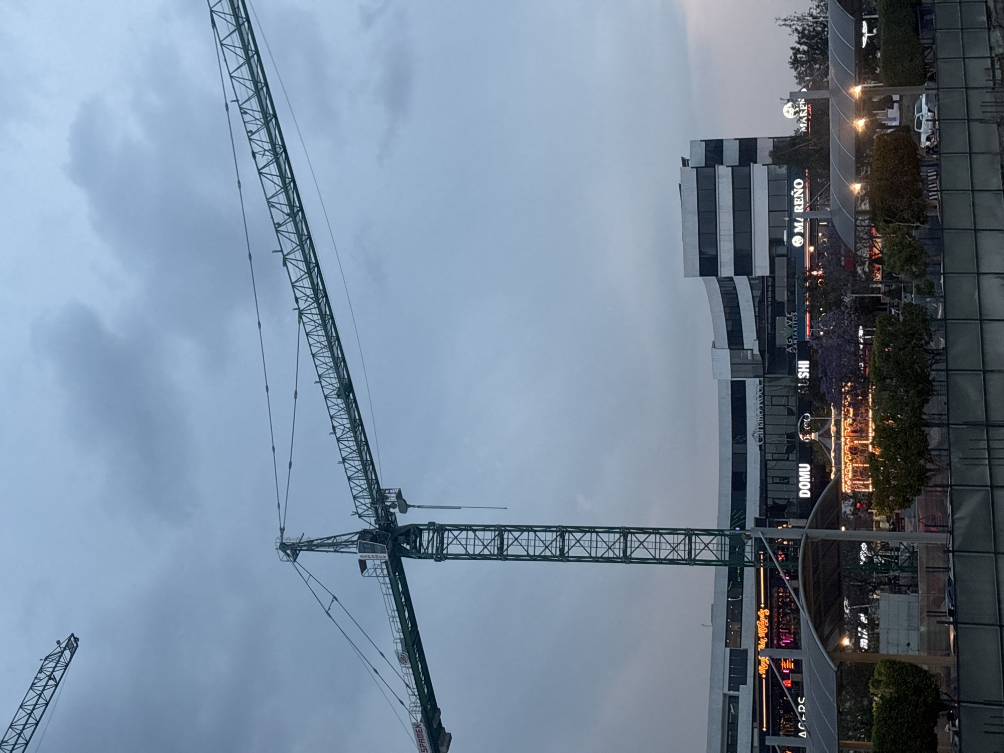 The image shows a construction site in Puebla, Mexico. There are two cranes in the image. The crane in the center is taller, and a shorter crane is partially visible in the lower left corner. The sky is cloudy. The city buildings are on the right side of the image, including the "MARENO" building. Other signs such as "DOMU" and "AGAPE CANTARTITOS" are also visible.