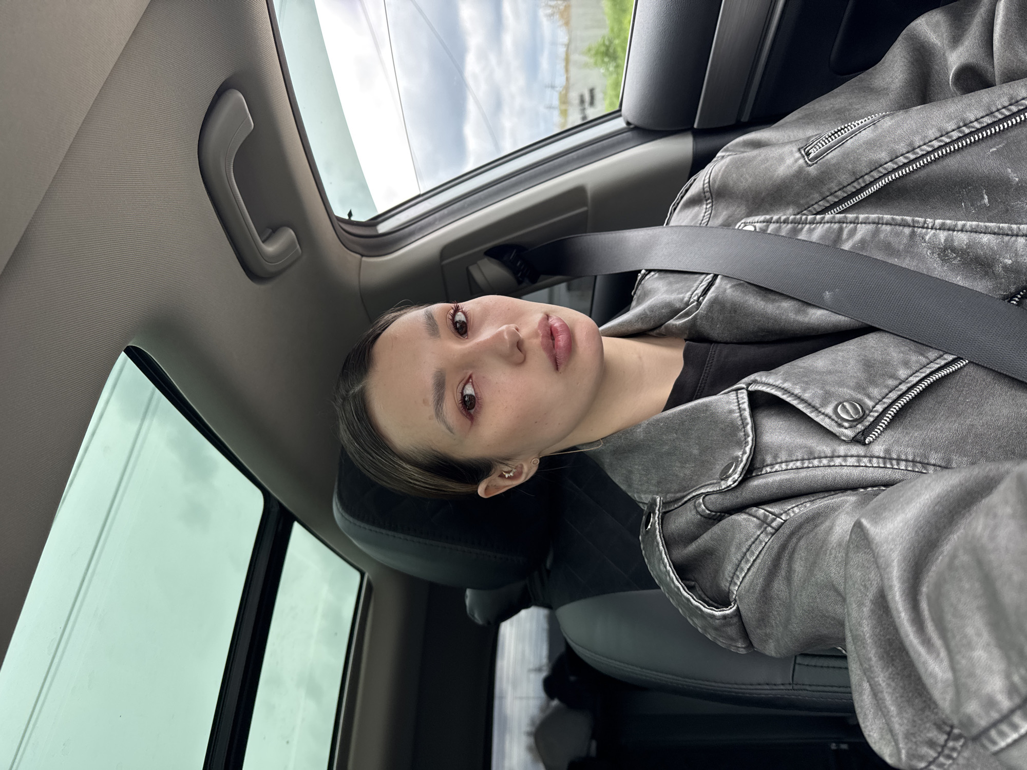 A medium shot taken from within a car, facing a person wearing a gray jacket with a seatbelt. The person has light skin and dark hair with light streaks. A car window is visible with clouds and green foliage outside. The car's interior is gray.