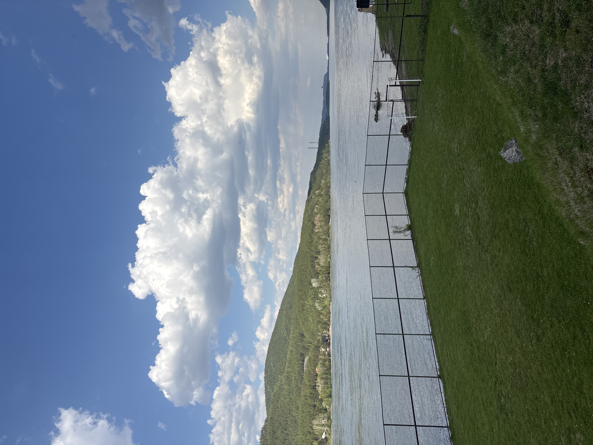 The image shows a body of water, likely a river, with a forested hill on the opposite bank. The sky is blue with large, puffy white clouds. In the foreground, there is a grassy area, possibly a riverbank, with a wire fence. The reflection of the fence is visible on the water. The scene could be in Овсянка, Russia, given the landscape features (river, forested hills) which are consistent with the region's geography.