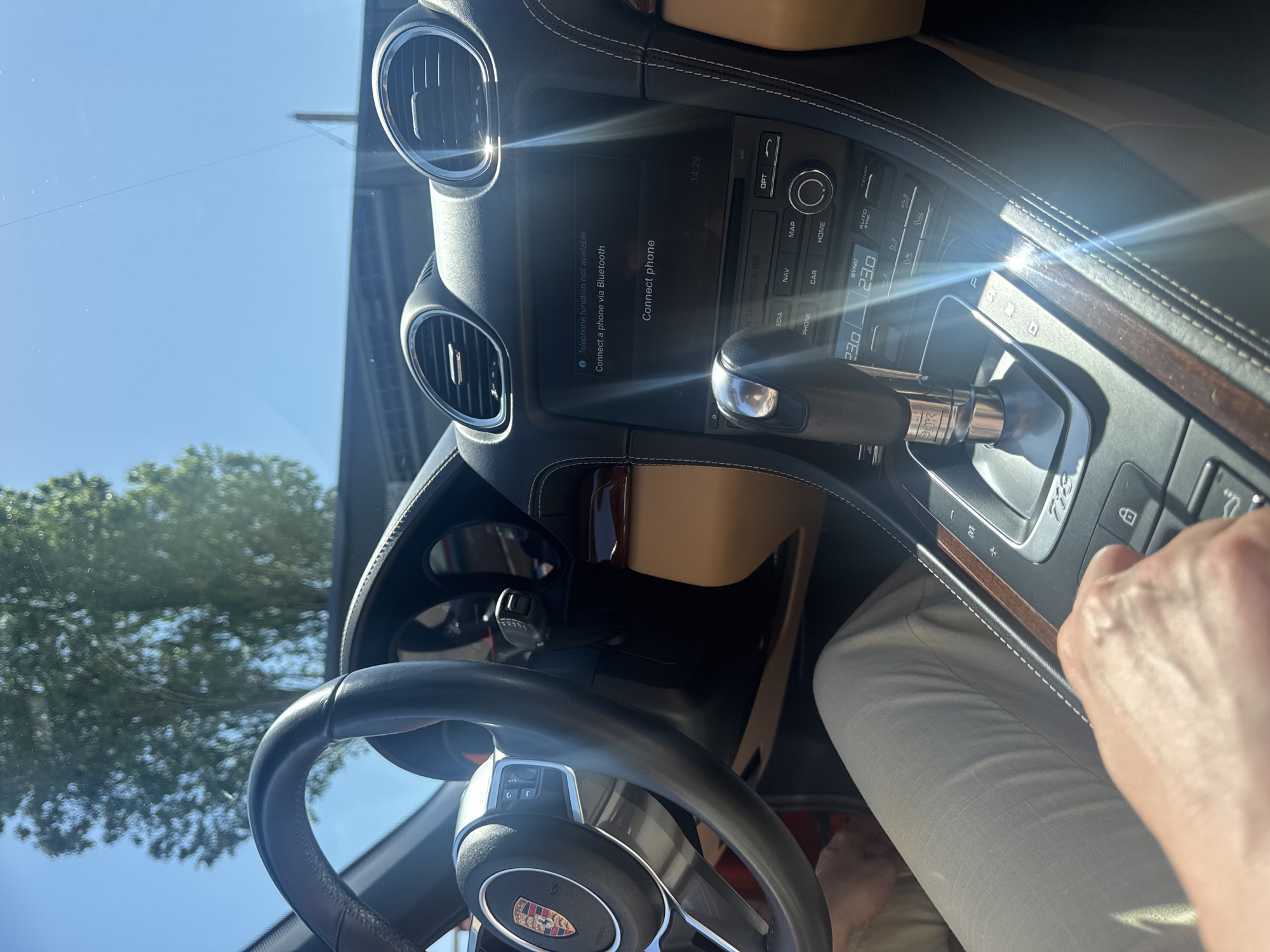 The image shows the interior of a Porsche, specifically the driver's side. The steering wheel, dashboard, and center console are visible. The infotainment system displays a message "Connect phone" with options to connect via Bluetooth. The temperature is set to 23.0 degrees. A hand is visible near the gear selector. The environment outside the car shows trees and a blue sky. It is impossible to tell if the location is in Sochi, Russia.