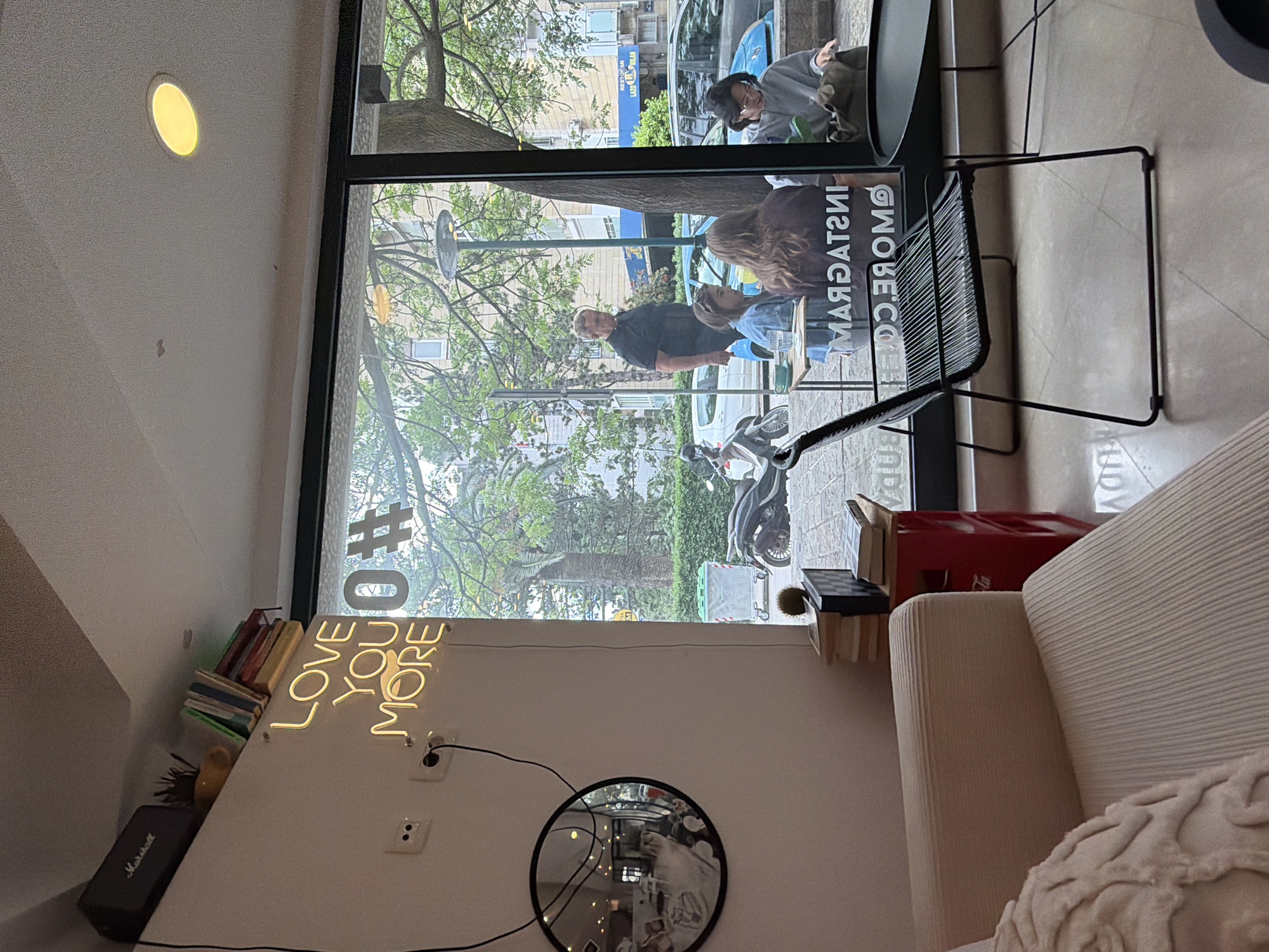 This is an indoor shot of a cafe interior in Budva, Montenegro. A window displays a street scene with pedestrians, parked cars, and a motorbike. Reflected in the window are cafe patrons at an outdoor table. Indoors, a "Love You More" neon sign is attached to the wall, along with books, a speaker, and a round mirror. A chair sits near the window and a portion of a sofa is in the foreground. The window also shows the cafe's branding which is “INSTAGRAM.MORE.CO”.