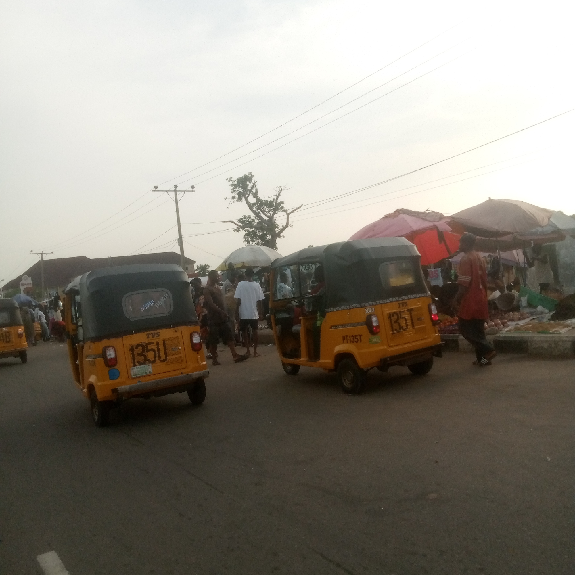 The image depicts an outdoor street scene in what appears to be a market area. In the foreground, two yellow auto-rickshaws (also known as tuk-tuks) are visible. The auto-rickshaw on the right is closer and more detailed, showing a license plate with the characters "PT135T". It has a black roof and a colorful patterned border along the bottom of the body. The auto-rickshaw on the left is further away and partially obscured, with its license plate showing "135U".
The background shows a bustling street with several people. Some individuals are walking or standing near market stalls, which are covered by umbrellas and awnings of various colors, including red and pink. One auto-rickshaw is parked or driving slowly behind the first one on the left. The environment suggests a commercial area, possibly a street market. Power lines and utility poles are visible against the overcast sky, indicating an urban setting. A bare tree branch with some leaves is also visible in the mid-ground. The overall lighting suggests it is daytime, with diffused light from the cloudy sky.