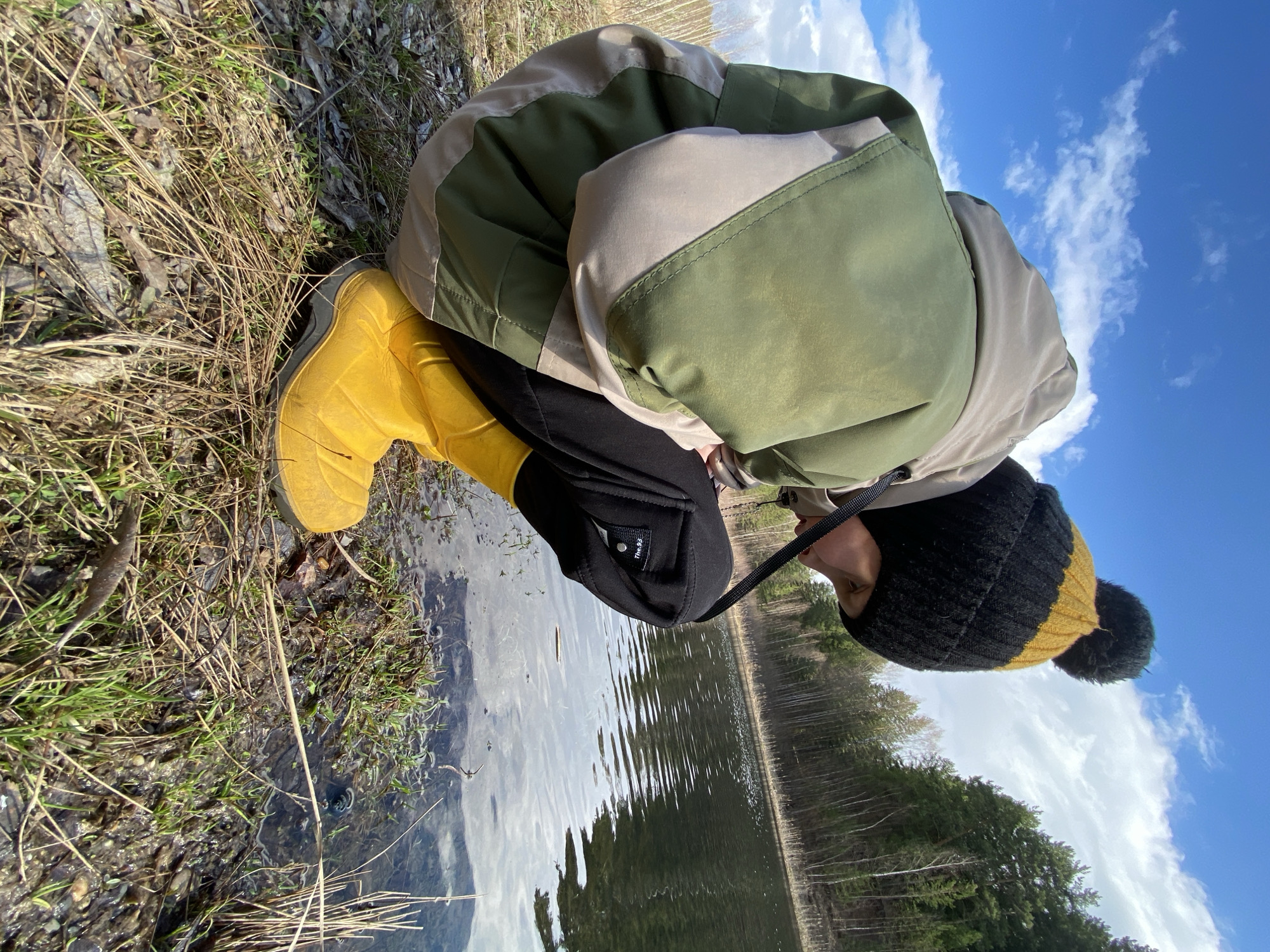 A person is crouched near the edge of a body of water. The person is wearing a green and beige jacket, black pants, yellow rubber boots, and a black and yellow hat with a pom-pom. The person is looking down at the water. The background shows trees and a blue sky with white clouds. The location is in the city of Noviy, Russia.