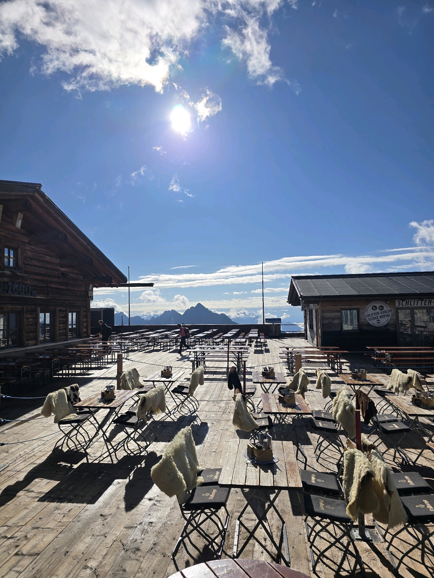 The image depicts an outdoor seating area of a mountain restaurant or hut, likely at a high altitude, given the mountainous backdrop and the clear blue sky with scattered clouds. The setting appears to be in or around Garmisch-Partenkirchen, Germany, a region known for its alpine scenery and ski resorts.
In the foreground, there are numerous wooden tables and chairs arranged on a wooden deck. Many of the chairs are draped with sheepskin or wool throws, suggesting a cooler climate. The tables are mostly empty, with a few small baskets or containers on them. The sun is bright, casting long shadows across the deck, indicating it is likely midday.
To the left, a rustic wooden building with a pitched roof and visible timber construction is present. Text that appears to read "2600 m" is visible on the building's facade, reinforcing the high-altitude location. To the right, another wooden structure with a dark roof houses what seems to be a service counter or shop, as indicated by a circular sign on the wall that includes text like "SCHLITTEN-VERLEIH" (Sledge Rental).
In the mid-ground, several people are visible. One person is standing near the wooden building on the left, appearing to interact with the tables and chairs. Another person, wearing a backpack, is walking across the deck on the right side, in the vicinity of the second building. Further back, a long row of empty picnic tables and benches stretches into the distance, set against a panoramic view of mountains under a bright sky. The overall impression is that of a serene, picturesque, and well-equipped mountain establishment ready for patrons.