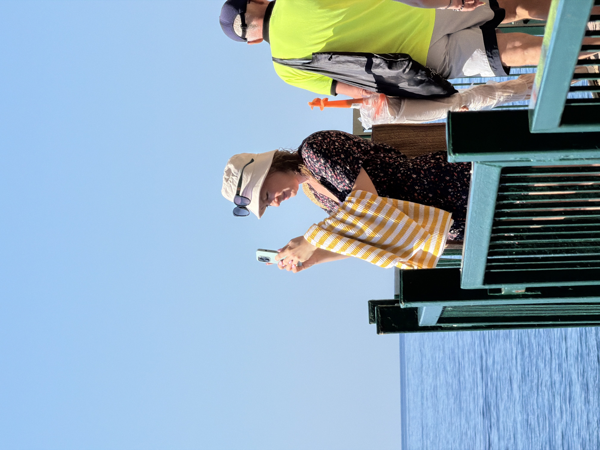 The image depicts a sunny day at a waterfront location in Budva, Montenegro. A woman wearing a floral print dress and a bucket hat stands on a wooden pier, holding a smartphone in her right hand and a yellow and white striped towel in her left. She appears to be taking a photo or video with her phone. To her right, a man wearing a neon yellow t-shirt and a baseball cap stands with his back to the camera, a black bag slung over his shoulder. In the background, the calm blue water of the sea stretches to the horizon under a clear sky. A green railing runs along the edge of the pier. Several objects are visible on the pier near the woman and man, including what appears to be an orange object and some white plastic wrapping. The overall atmosphere is that of a relaxed outdoor excursion.