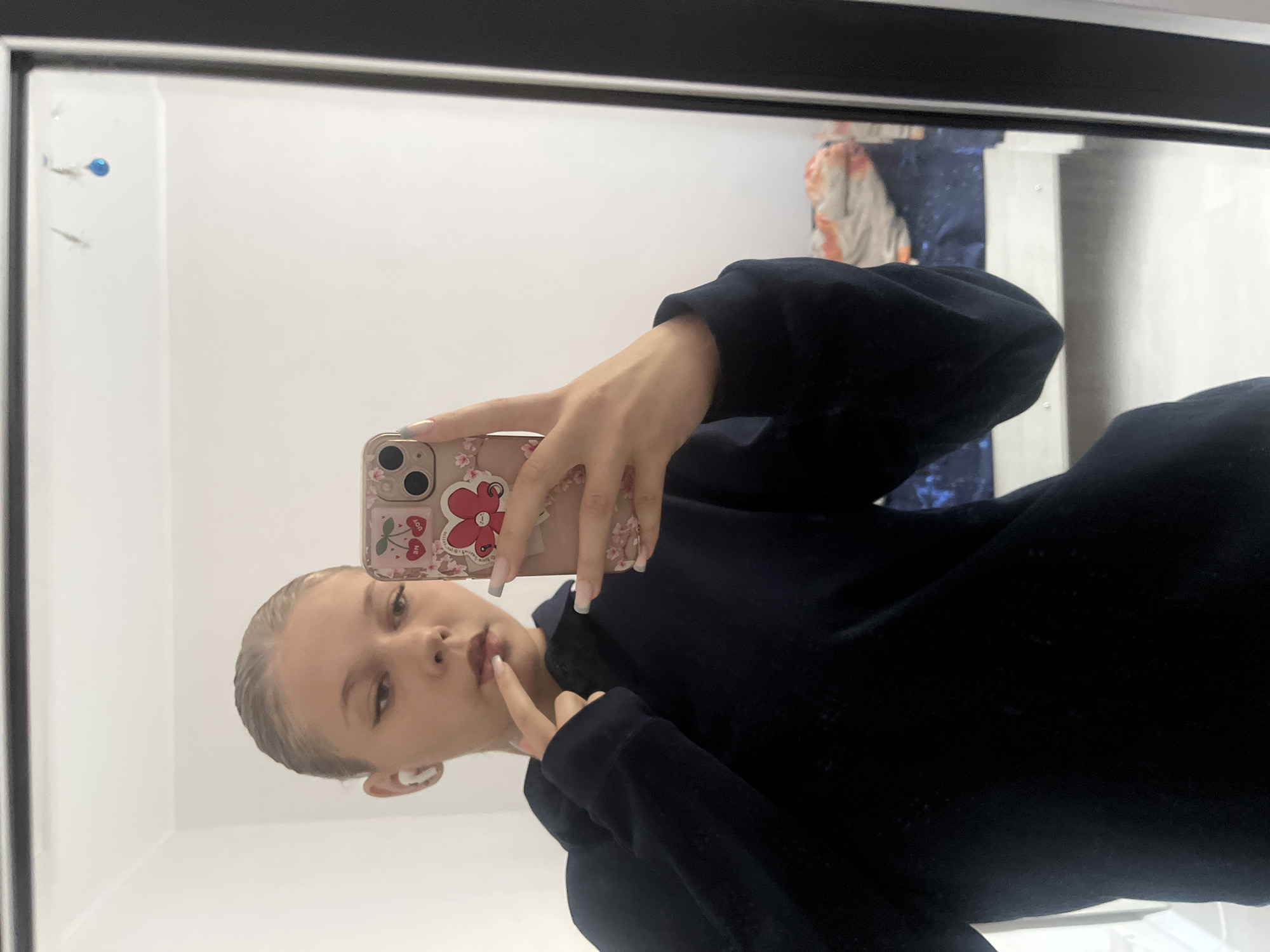 A young woman with blonde hair pulled back is taking a mirror selfie in what appears to be a bedroom. She is wearing a dark blue hoodie and has white wireless earbuds in her ears. Her left hand is touching her chin and lips, and her right hand is holding a smartphone with a pink case featuring cherry and floral designs. The background of the reflection shows a white wall, a dark framed mirror, and part of a bed with blue and orange bedding. The lighting suggests an indoor environment. There is no identifiable information about the location in the city of Perm, Russia.