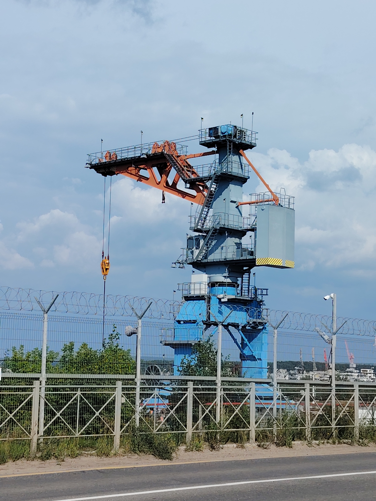 The image shows a large industrial crane with an orange boom and a blue tower, situated in an industrial area. The crane is equipped with a hook hanging from a cable. In the foreground, there is a fence with barbed wire and a CCTV camera mounted on a pole. Behind the fence, there are green trees and bushes. In the distance, a cityscape with other industrial structures and cranes is visible under a cloudy blue sky. The location is the port of Chaikovsky, Russia.