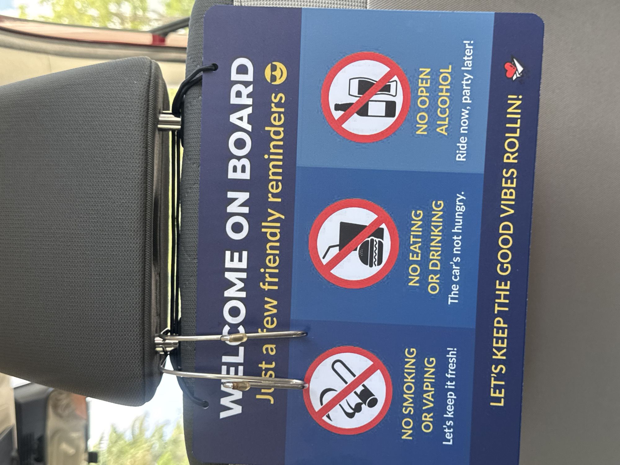 This is a vertical close-up shot of a car's interior, specifically a sign hanging from the driver's side headrest. The sign is dark blue with yellow text and features three red-circled icons with diagonal red lines through them. The first icon depicts an open bottle and a can, with the text "NO OPEN ALCOHOL Ride now, party later!" written next to it. The second icon shows a burger and fries, and the accompanying text reads "NO EATING OR DRINKING The car's not hungry." The third icon features a cigarette and a vape pen, with the text "NO SMOKING OR VAPING Let's keep it fresh!" below it. The sign also has the heading "WELCOME ON BOARD Just a few friendly reminders." At the bottom, it says "LET'S KEEP THE GOOD VIBES ROLLIN!" with a small red heart and paper airplane icon. The scene appears to be daytime, with natural light filtering in from outside, suggesting it's a sunny day. The focus is on the sign, but the dark grey fabric of the headrest and the edge of the car's window are also visible. The overall tone of the sign is lighthearted and informative, aiming to set expectations for passengers. The location is identified as Aventura, United States of America.