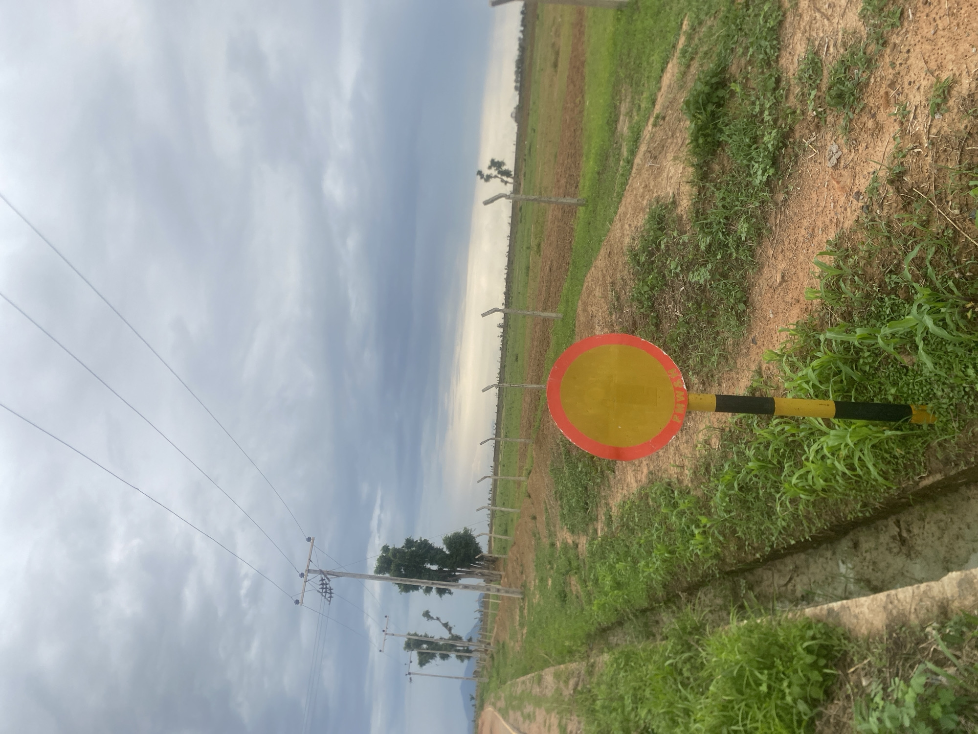 The image depicts an outdoor scene in Bulamare, Nigeria, under a cloudy sky, suggesting it is daytime. The perspective is from a low angle, looking upwards and towards a distant horizon. The foreground features a red and yellow circular sign with black stripes on its pole, positioned on the right side of the frame. The ground is a mix of dry soil and patches of green grass.
To the left, power lines stretch across the sky, supported by wooden utility poles. Beyond this, a fence runs along a field that transitions from cultivated patches of brown soil to areas of green vegetation. In the mid-ground, a few scattered trees stand with sparse foliage.
The overall setting appears to be a rural or undeveloped area, possibly on the outskirts of a town or village. The presence of power lines and agricultural fields indicates human activity, though no people are visible in the image. The cloudy weather creates a diffused light, and there is no visible text in the image. The scene evokes a sense of openness and a slightly overcast atmosphere.