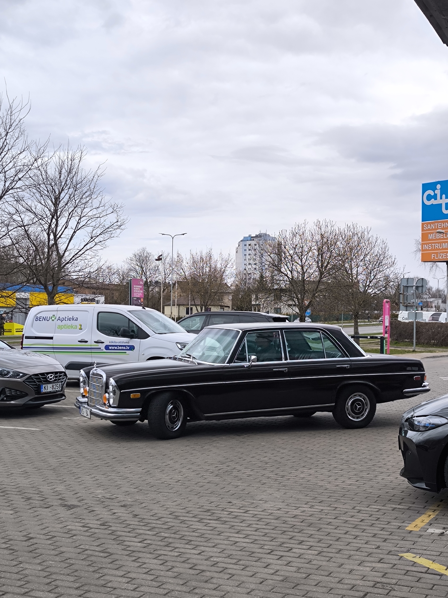 The image captures a cloudy day scene in Riga, Latvia, featuring a black classic Mercedes-Benz parked on a brick-paved parking lot. Adjacent to the Mercedes are other vehicles, including a gray Hyundai and a white BENU pharmacy van. A building and trees are visible in the background. In the upper right corner, there is a sign for a construction and furnishing supply business.