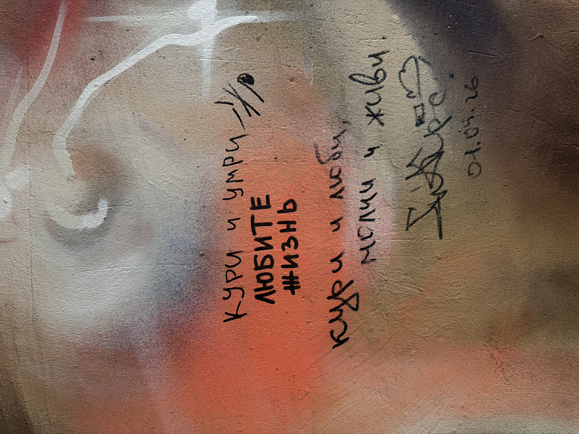 This is a photograph of graffiti on a textured wall. The graffiti is in black marker and appears to be handwritten. The text is in Russian and translates to "smoke and die / love life" on the top line and "smoke and love, be silent and live" on the bottom line. There is also a date written below the graffiti, which reads "01.05.26". The wall has a spray-painted background in shades of orange, brown, and white. The style of the graffiti suggests it was created spontaneously and without formal planning. The overall impression is one of urban decay and artistic expression in a public space.