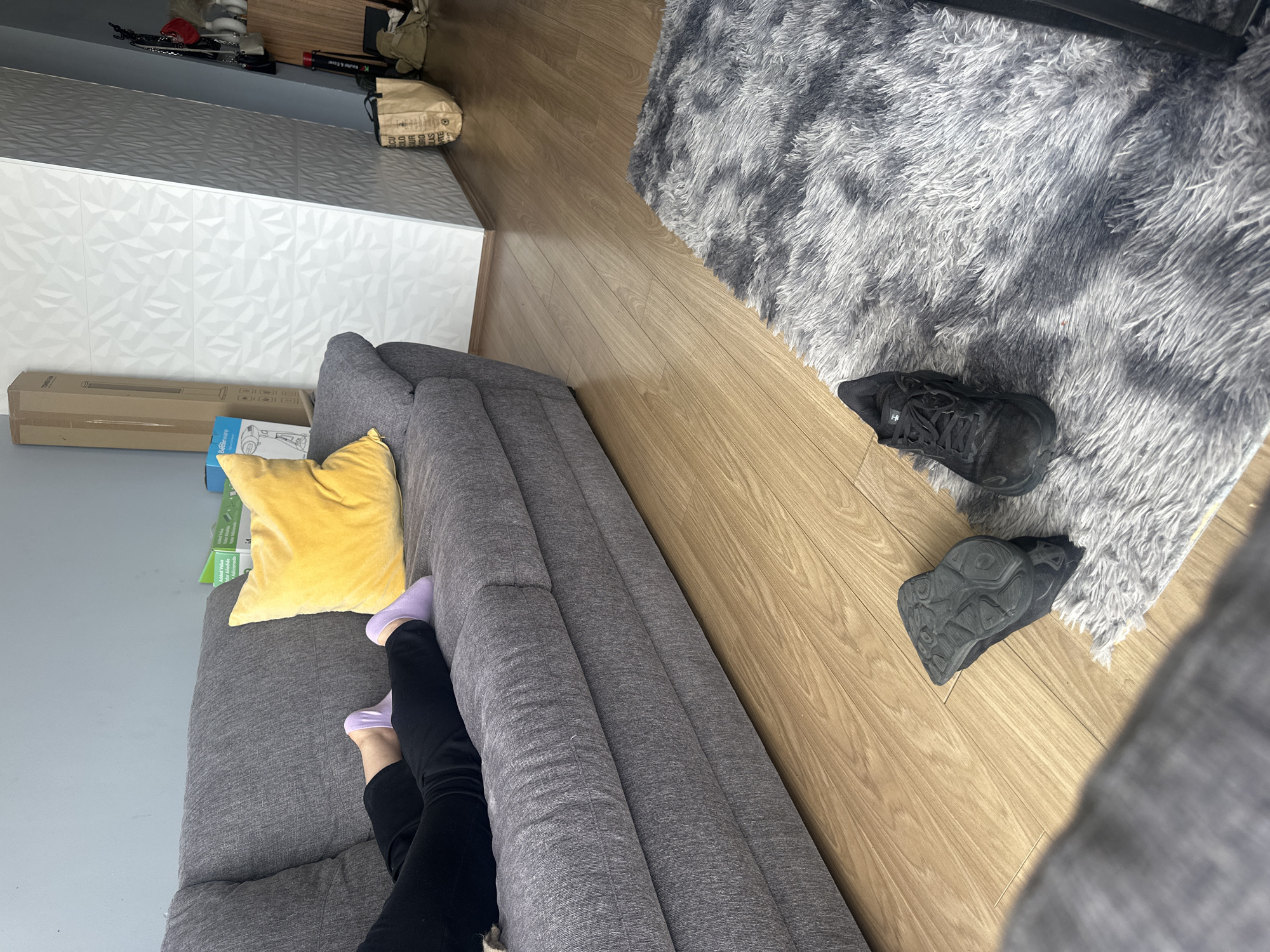 A high-angle shot shows the corner of a room, dominated by a gray sofa on the left. A person wearing black pants and lavender socks is reclining on the sofa. A yellow pillow rests on the sofa behind them. Next to the sofa, on a wooden floor, are two black shoes. A gray and white rug covers the floor to the right. In the background, a white cabinet and cardboard boxes are visible. The location appears to be indoors, potentially in a residence. Huixquilucan de Degollado, Mexico is not evident in the scene.