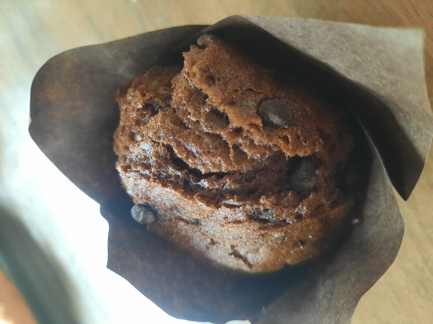 The image displays a close-up, overhead view of a chocolate muffin nestled within a crinkled brown paper wrapper. The muffin itself is a rich, dark brown color with a visibly moist and slightly irregular texture on its domed top, indicative of a baked good. Scattered across the surface and embedded within the muffin are several small, dark chocolate chips, adding visual interest and suggesting a rich flavor. The brown paper wrapper covers the lower part and sides of the muffin, with its upper edges softly folded or curled outwards. The background appears to be a light brown, possibly wooden, surface, with some lighter, almost white, areas on the left side, suggesting a light source. There are no people, explicit activities, specific location cues, weather conditions, emotions, or visible text in the image.