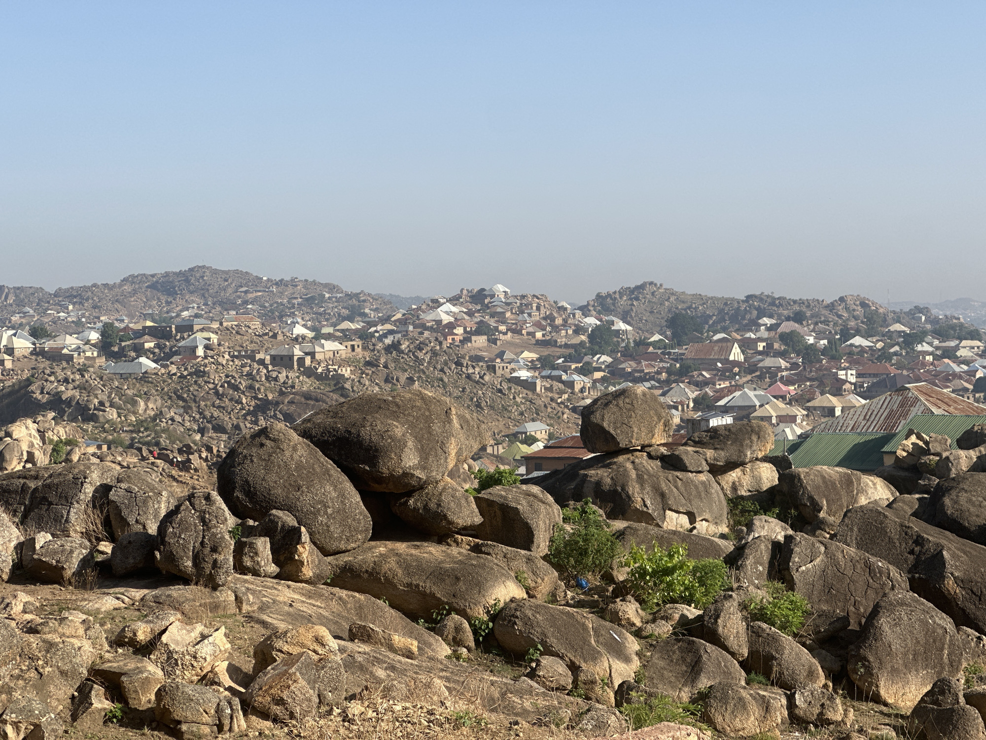 The image captures a section of Jos, Nigeria, viewed from a rocky outcrop. The foreground consists of large, weathered boulders interspersed with sparse vegetation. Behind the rocks, a dense settlement extends across a series of hills. The buildings are predominantly single-story structures with a mix of roof types and materials, including corrugated metal. The overall scene is bathed in a hazy, muted light under a clear sky.