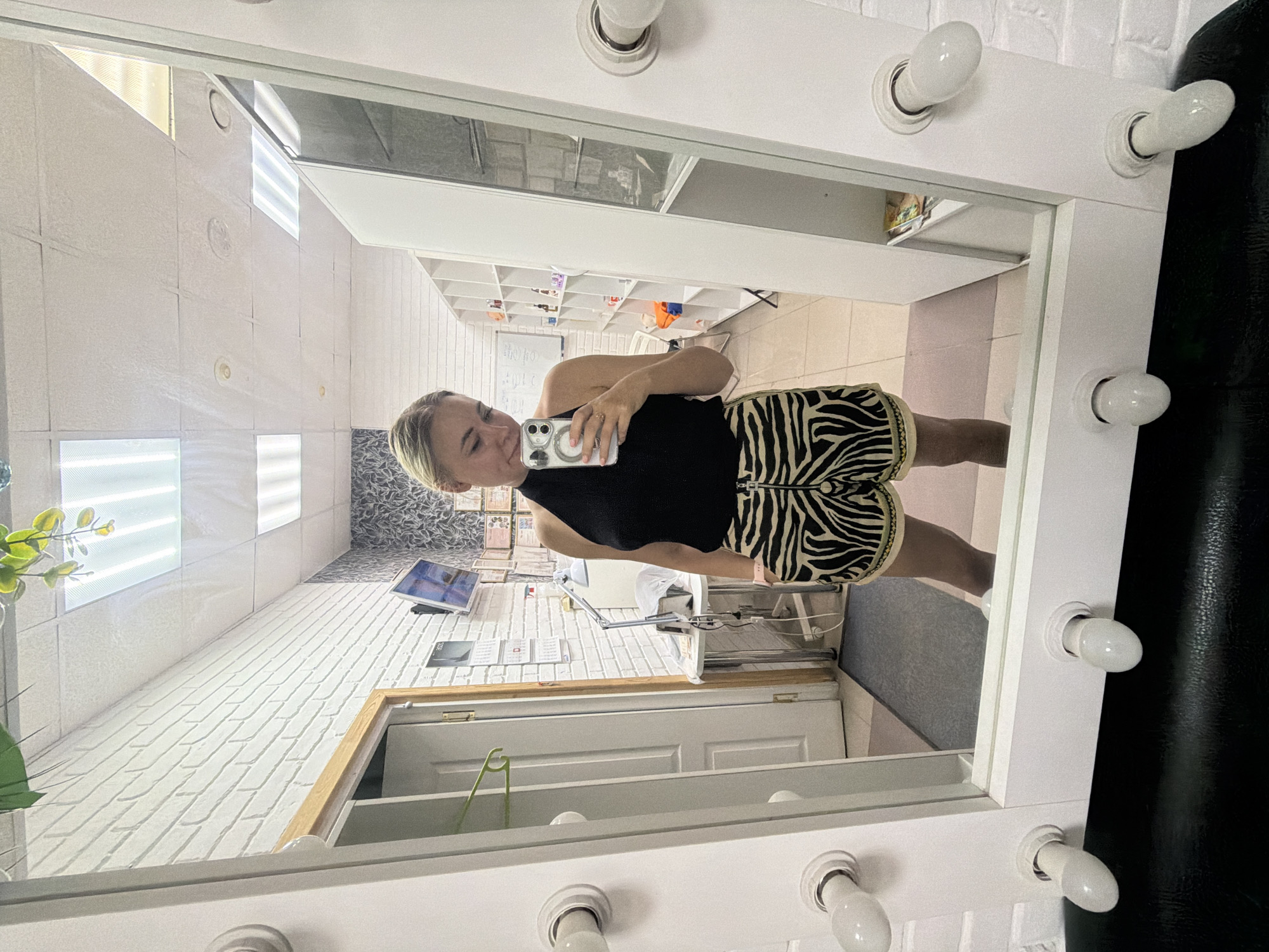 A woman stands in a room, reflected in a large, brightly lit mirror with bulbs around its edge. She is holding a smartphone in her right hand, taking a selfie. The woman is wearing a black halter top and zebra-print shorts. The room appears to be a salon or studio, with white brick walls, shelves filled with products, a desk with a lamp, and a door. The mirror is framed in white and has a black surface below it, possibly a chair. The scene is consistent with an interior setting in Chaikovsky, Russia.