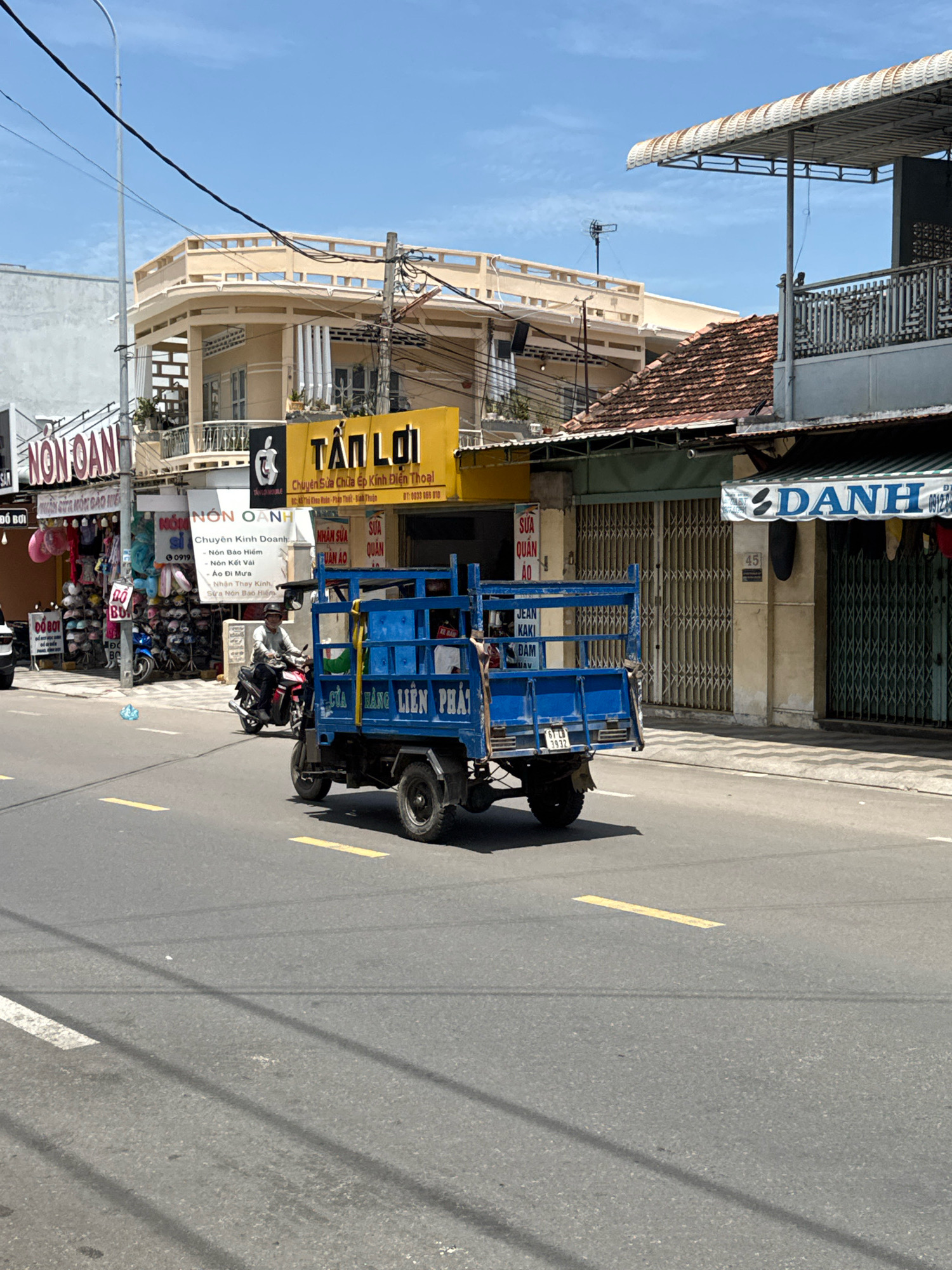 In Phú Thủy, Vietnam, a blue, three-wheeled truck with a cage-like rear cargo area is driving on a paved road under a clear blue sky. The truck has "LIÊN PHÁT" written in white on its side. A person wearing a helmet is driving the truck. The road has yellow lane markings and a white line on the left side. In the background, there are buildings with shops and balconies. One building has a yellow sign with Vietnamese text that includes "TÂN LỢI". Another sign reads "NON OANH". A motorcycle is parked to the left of the truck. The scene depicts a street in a commercial area.